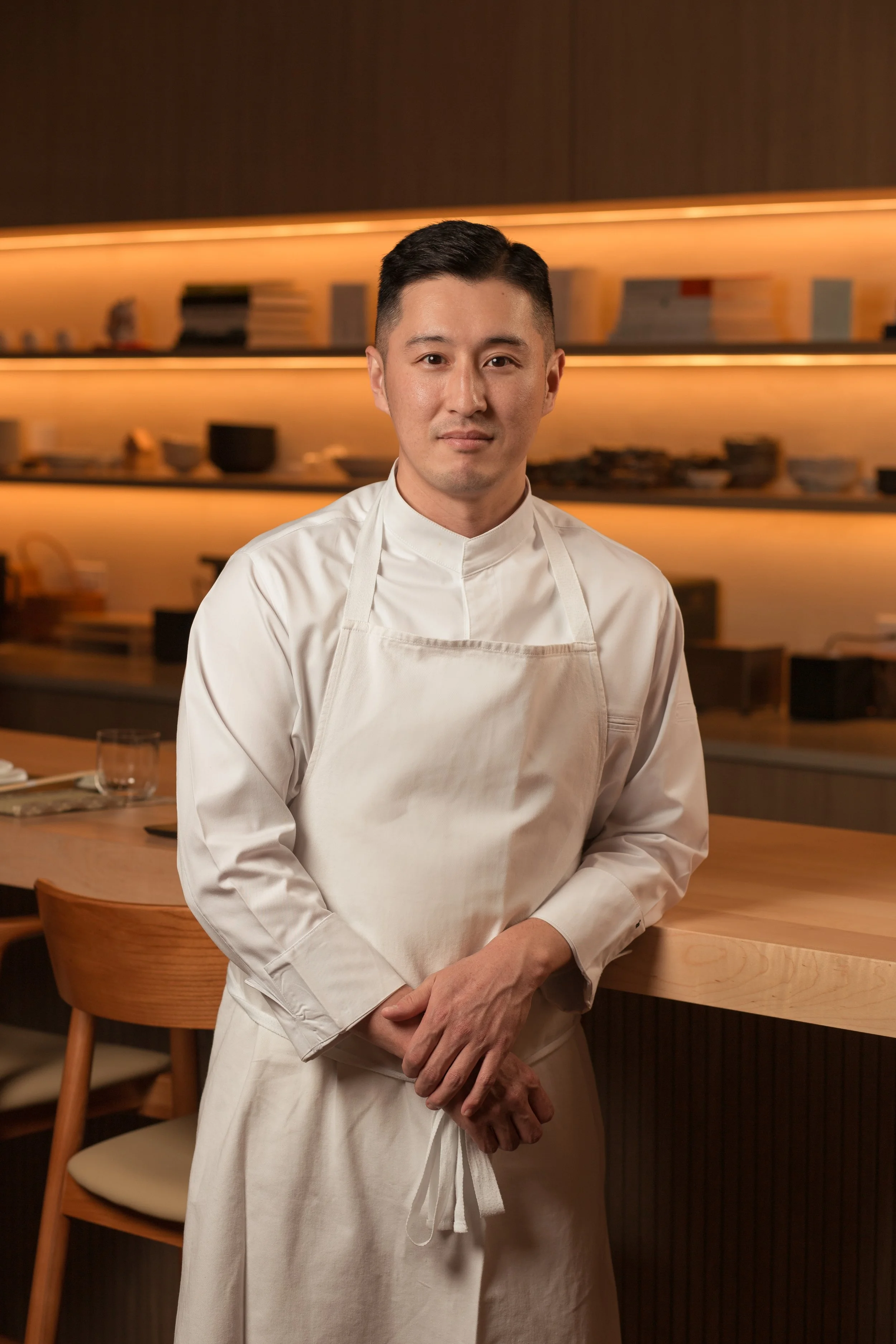 A male chef dressed in a white chef’s coat and apron standing in a restaurant kitchen or dining area, with wooden shelves and plates in the background.