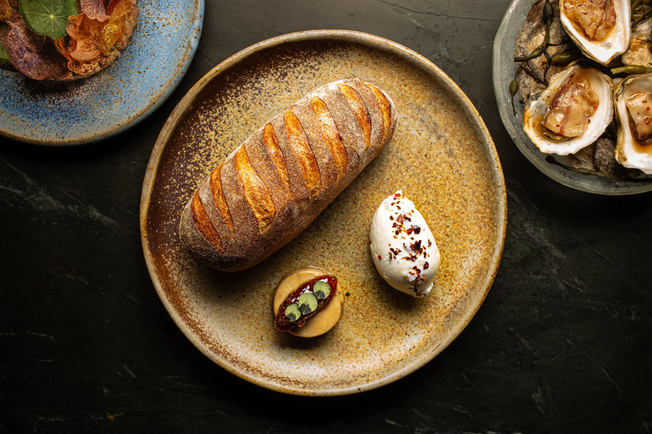 A ceramic plate with a loaf of baked bread and three different spreads or toppings, one garnished with herbs, on a dark surface. Surrounding dishes contain additional food items.