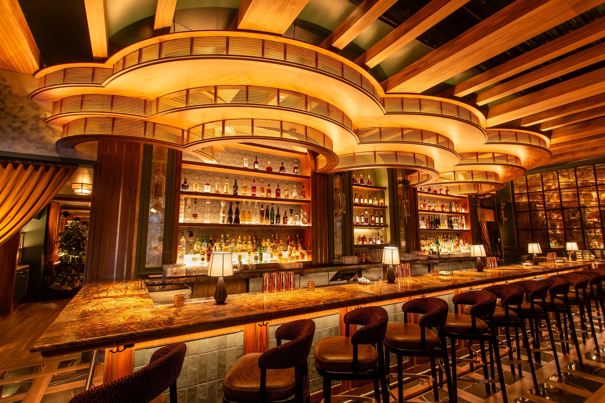Interior of a modern bar with a curved, illuminated wooden ceiling above a marble-topped counter, shelves with various bottles of alcohol, and bar stools along the counter.