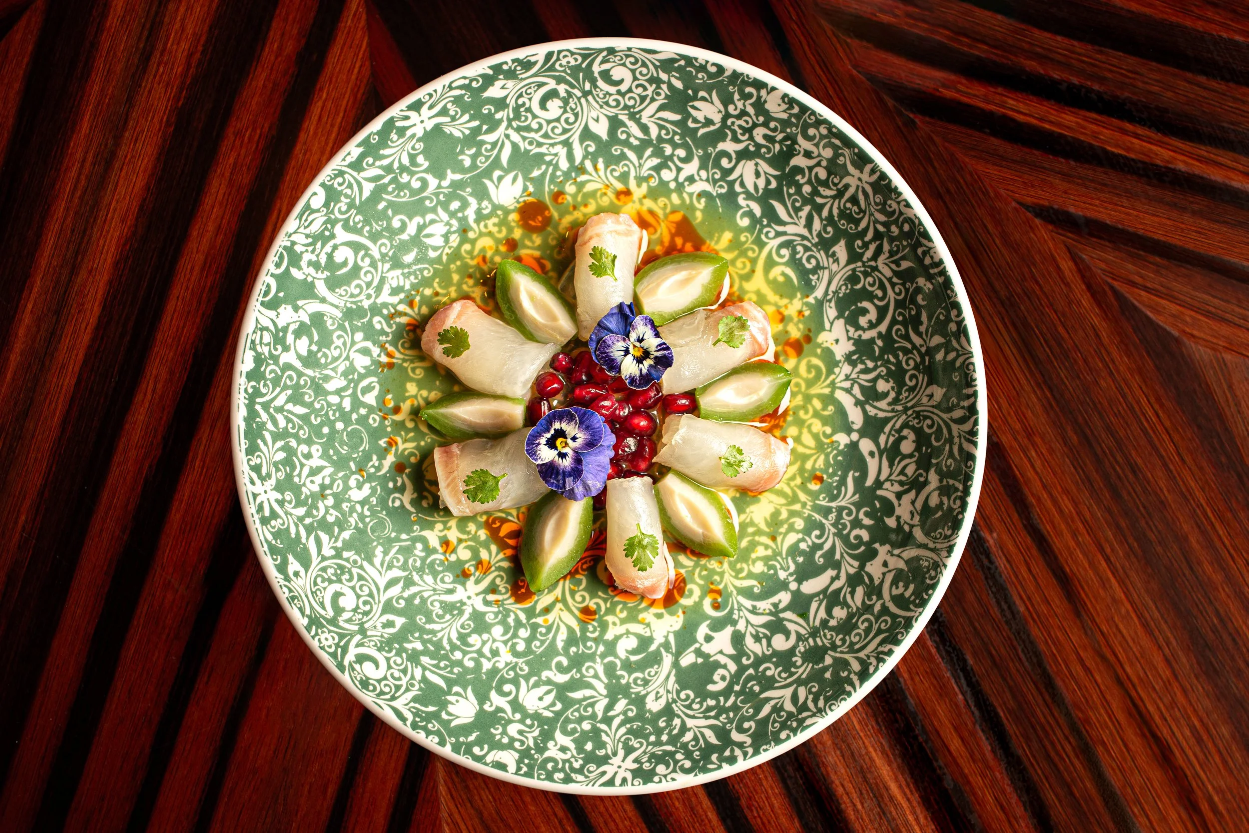 A decorated plate featuring slices of fish, garnished with herbs, edible flowers, and pomegranate seeds, on a dark wooden table.