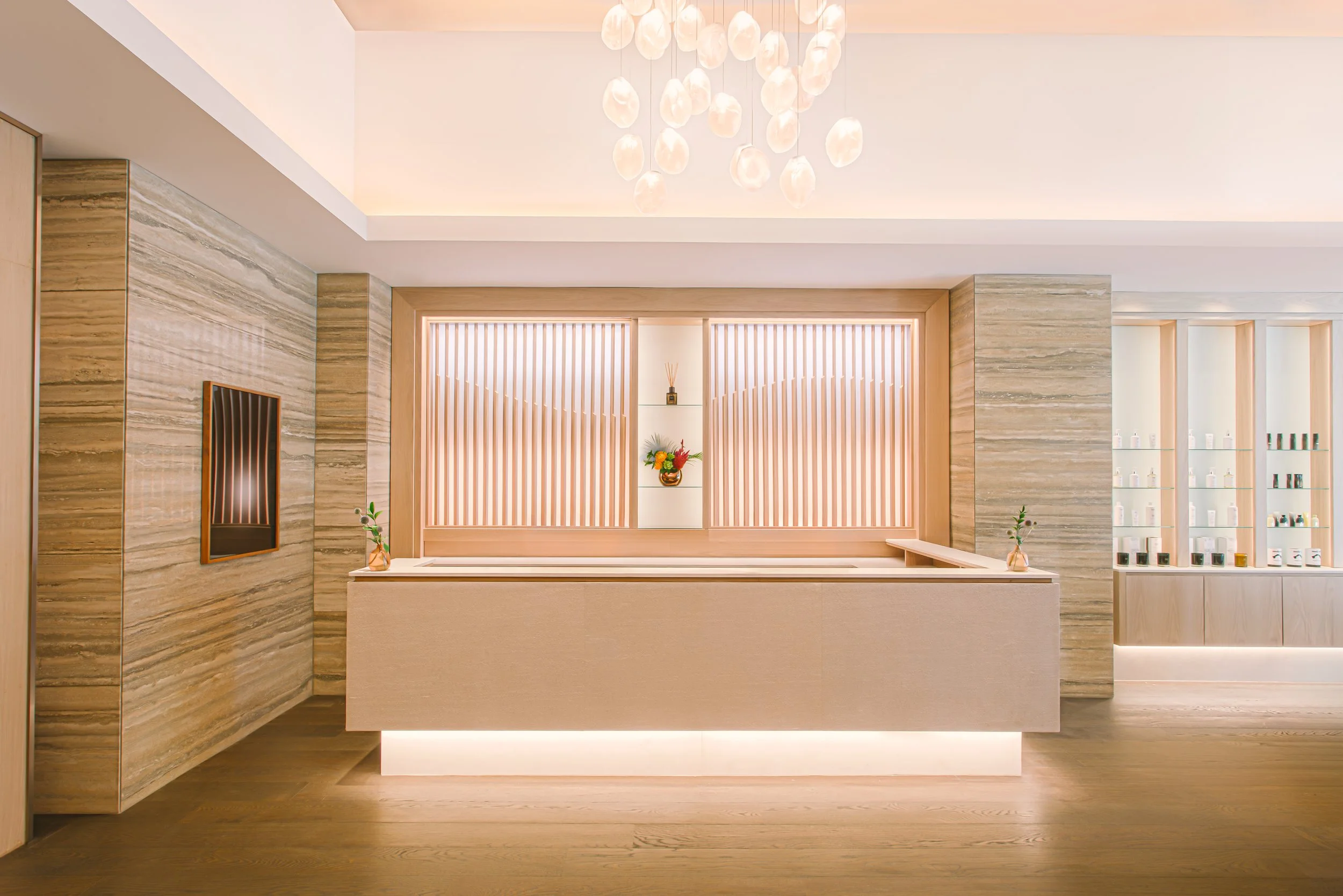 Modern reception area with beige and wood tones, decorative wall paneling, and display shelves with bottles, illuminated by soft lighting and a chandelier.
