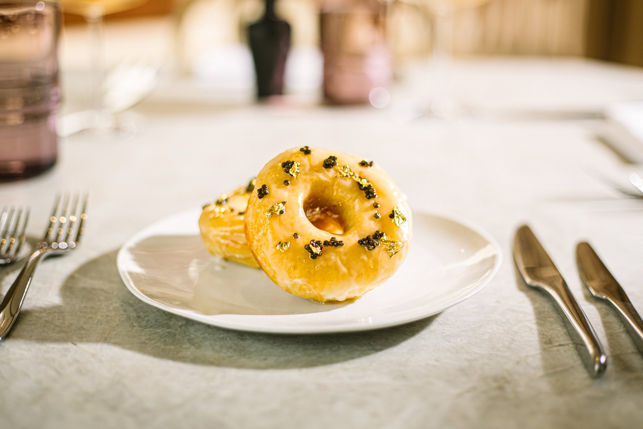 Two glazed donuts with black and gold sprinkles on a white plate on a table set for dining, with forks and knives visible.