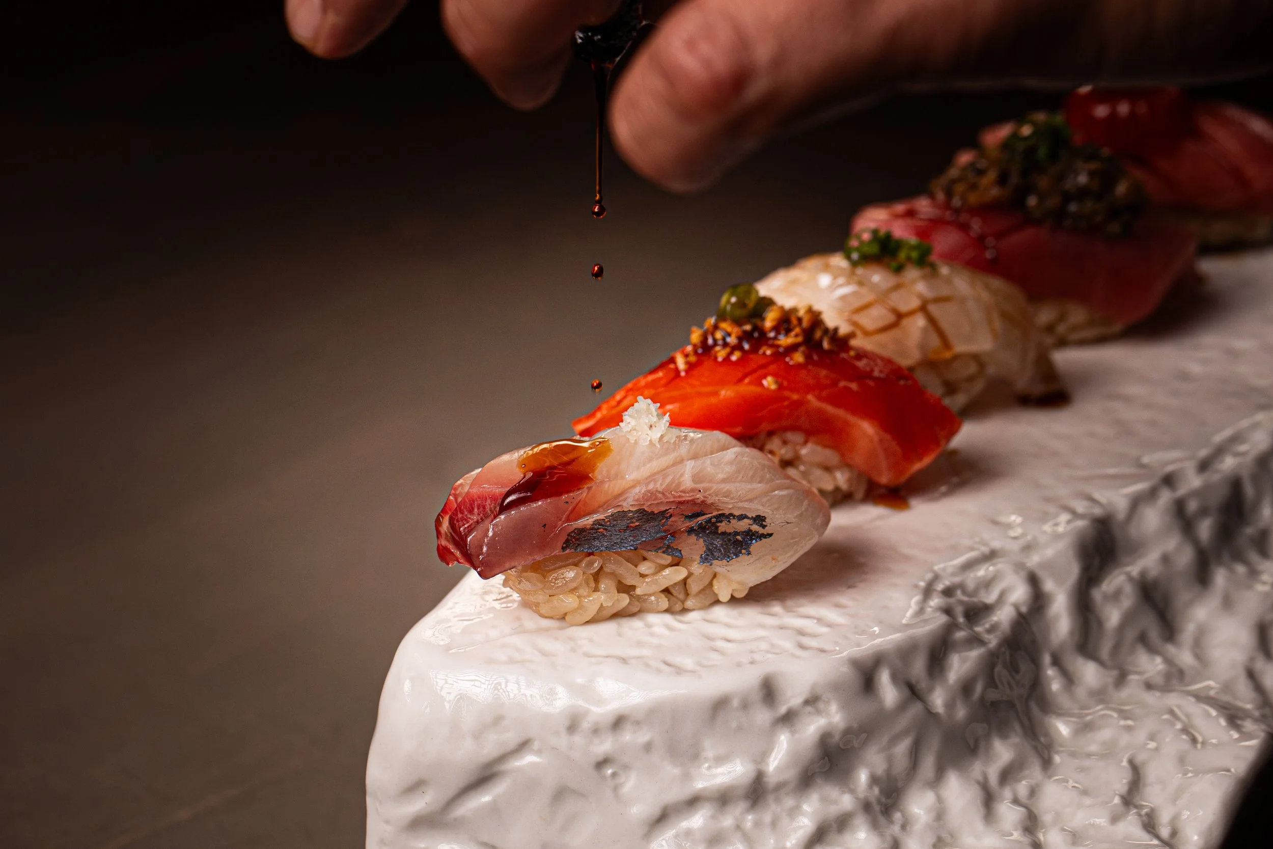 Close-up of a sushi platter with various raw fish pieces, garnished with sauces and toppings, being drizzled with soy sauce.