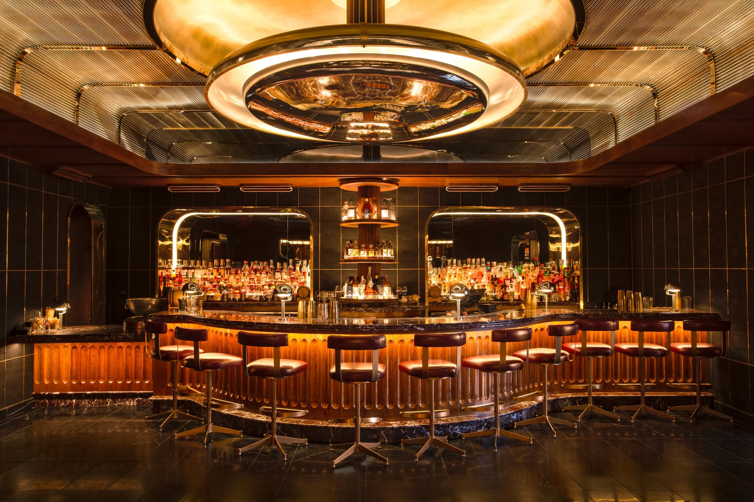 A luxurious bar with a curved marble countertop, leather bar stools, and a backlit display of various liquor bottles behind mirrors. The ceiling features modern metallic light fixtures, creating a warm, elegant ambiance.
