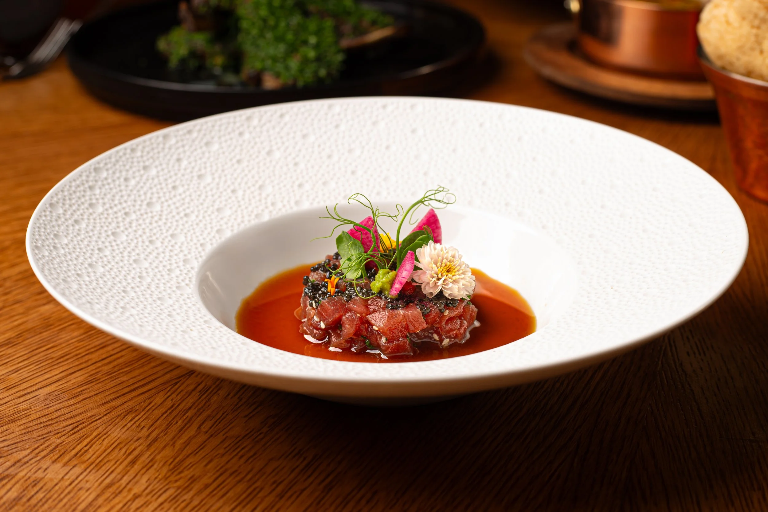 A white porcelain bowl with intricate textured patterns around the rim, featuring a raw tuna tartare garnished with microgreens, edible flowers, and thinly sliced vegetables, served in a shallow pool of sauce on a wooden table.