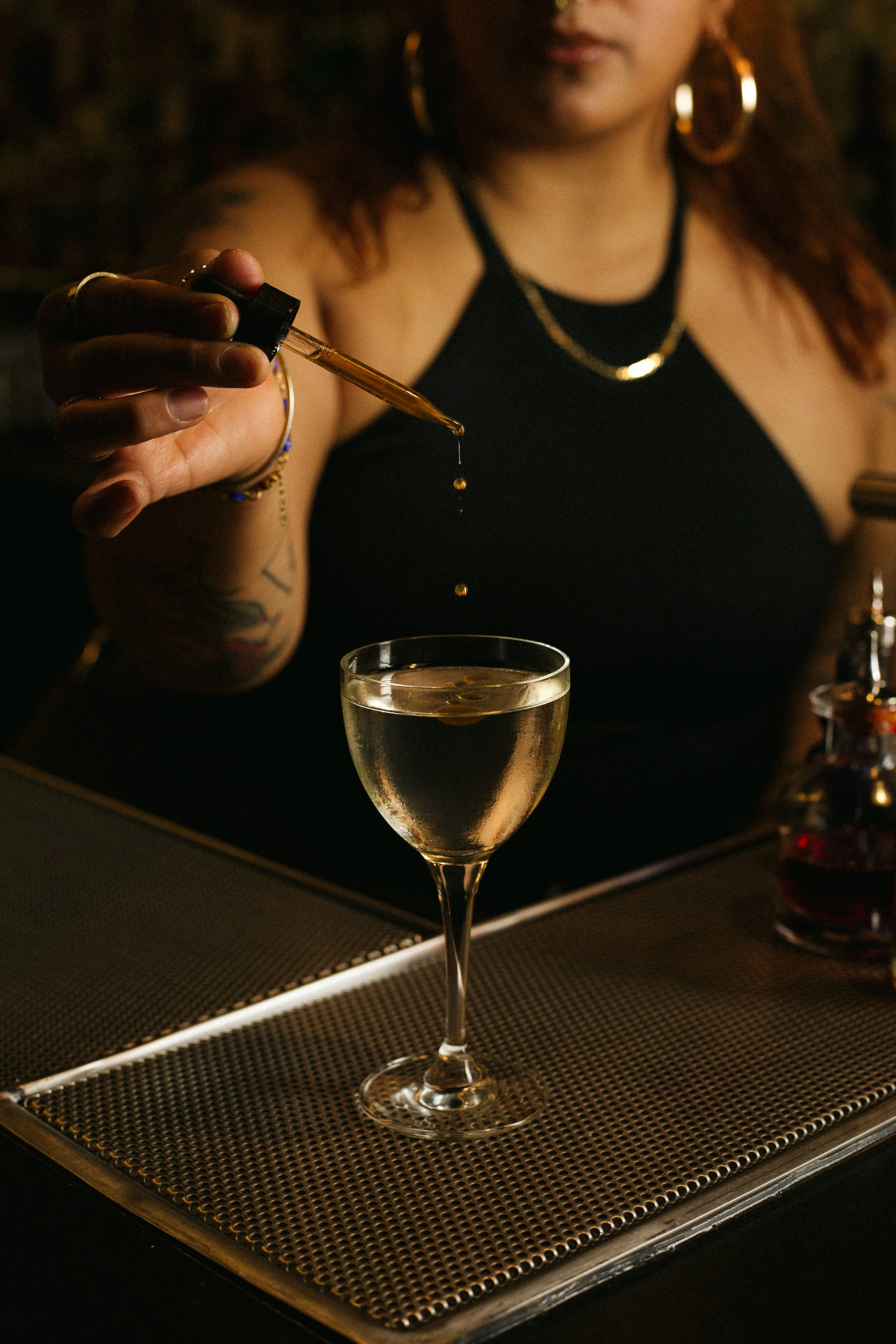 A woman with red hair and hoop earrings is dropping a liquid dropper into a glass of champagne at a bar.