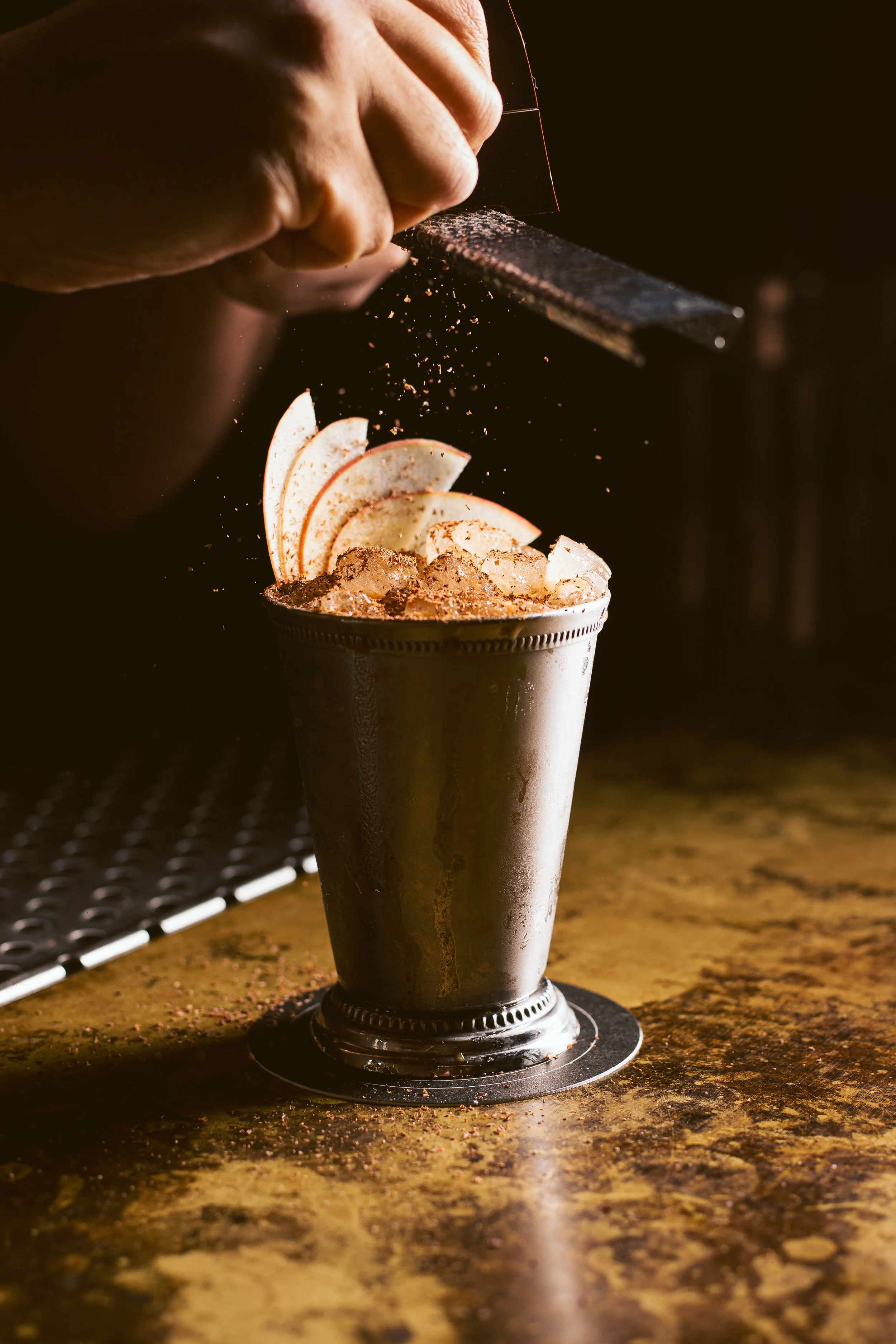 A bartender is grating nutmeg over a cocktail garnished with apple slices and ice in a metal cup.