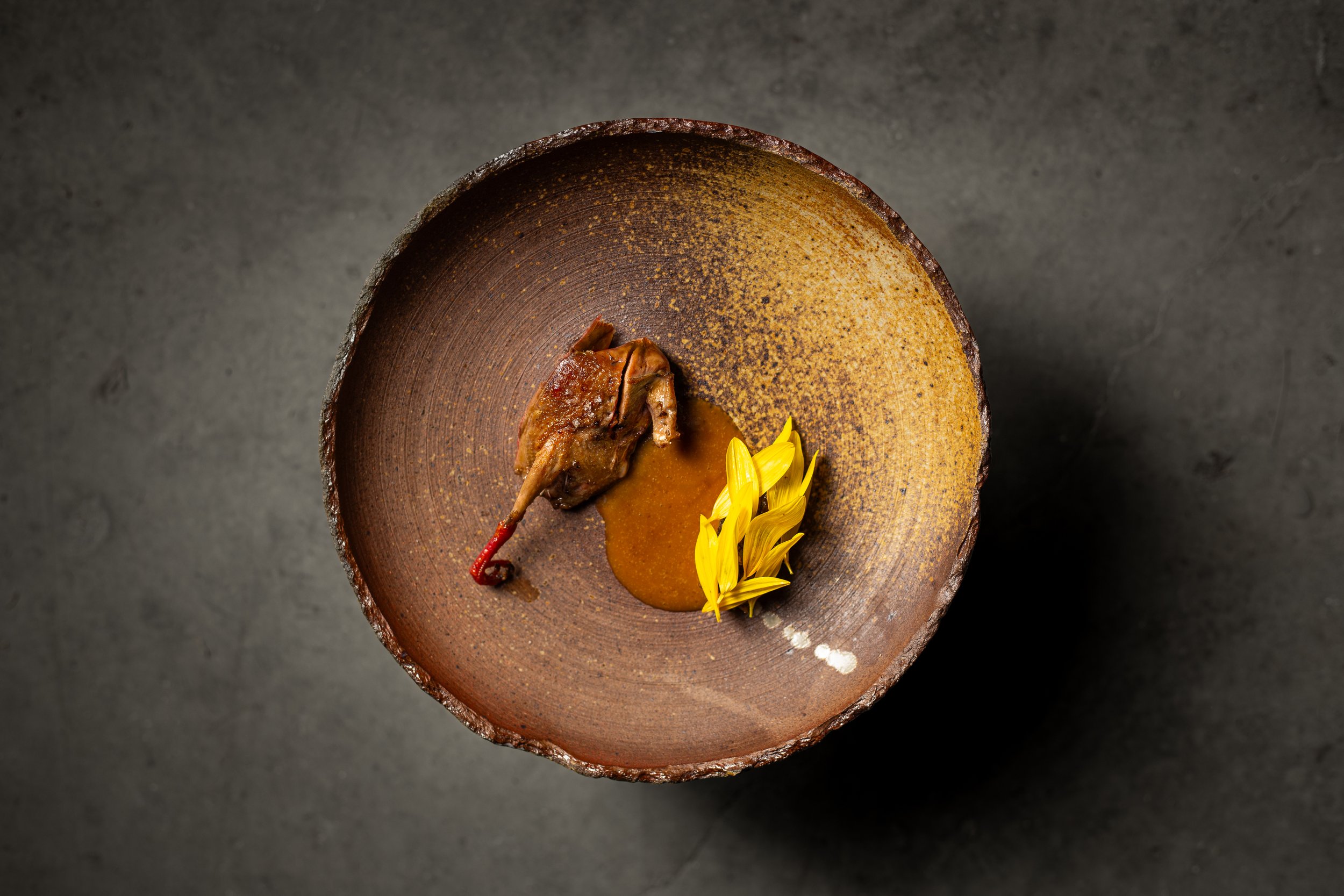 A brown ceramic bowl containing a small cooked bird, some sauce, and yellow flower petals, viewed from above.