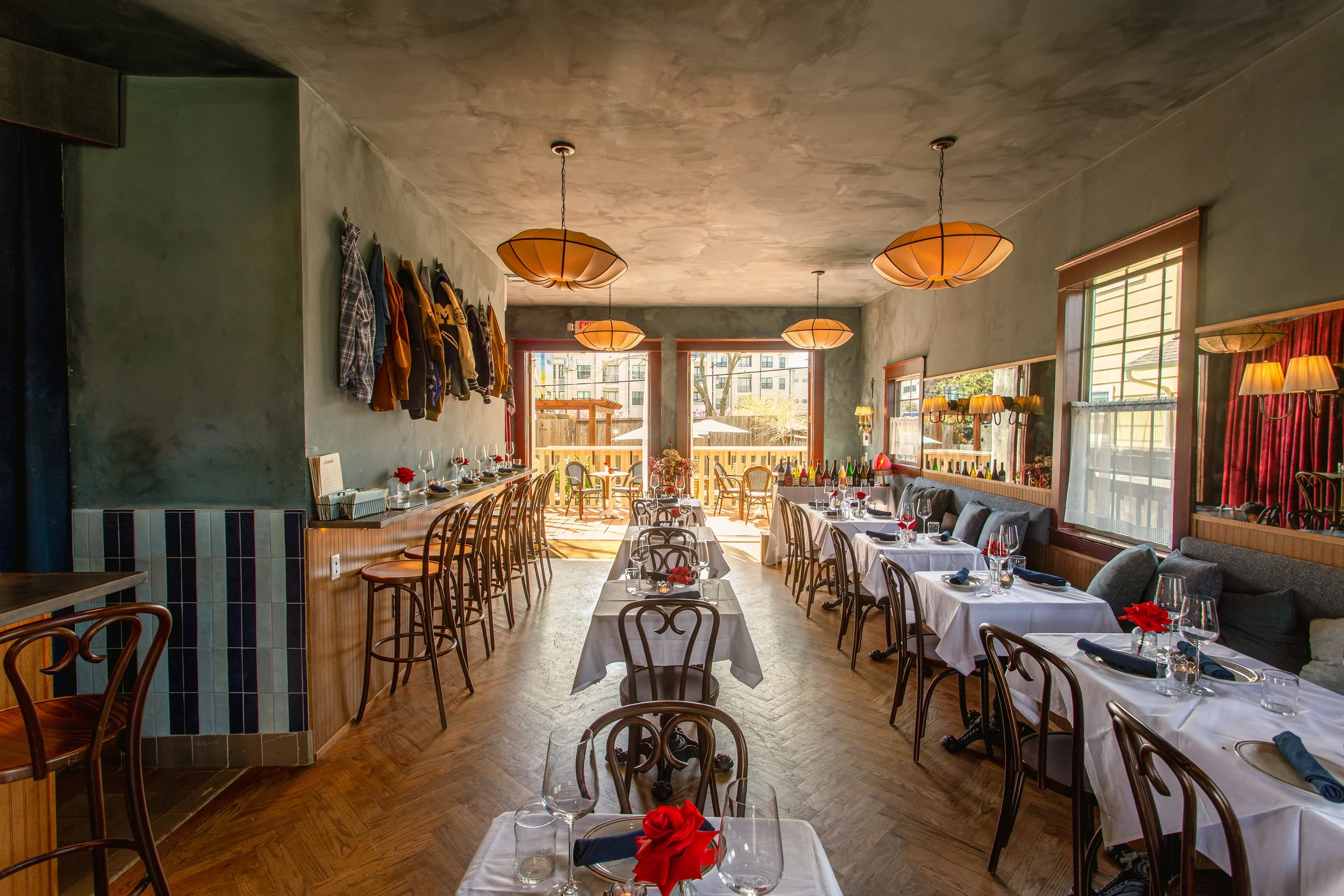A cozy restaurant interior with wooden floors, tables set with white tablecloths, red napkins, wine glasses, and flowers. The room has large windows allowing natural light, with hanging pendant lights and a gallery of jackets on the left wall. There is a patio area outside visible through the windows.
