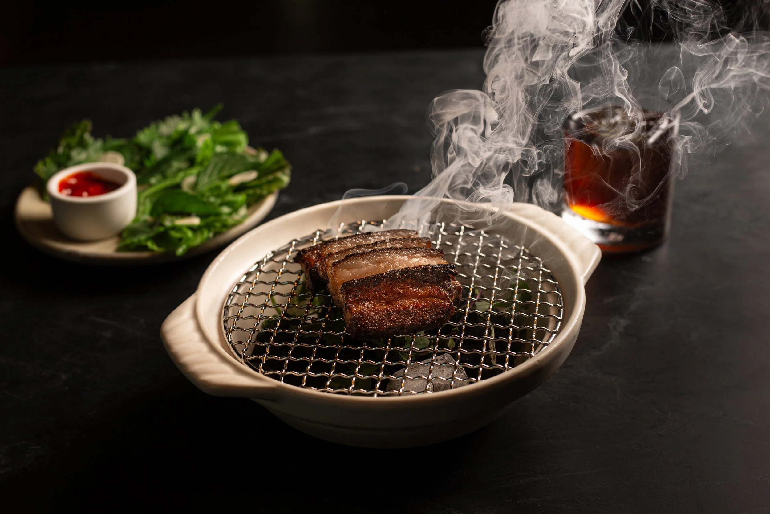 Sliced grilled meat on a wire rack over a ceramic grill, with smoke rising. In the background, a plate of fresh greens with a small bowl of red dipping sauce. A glass of dark beverage with ice on a dark surface.