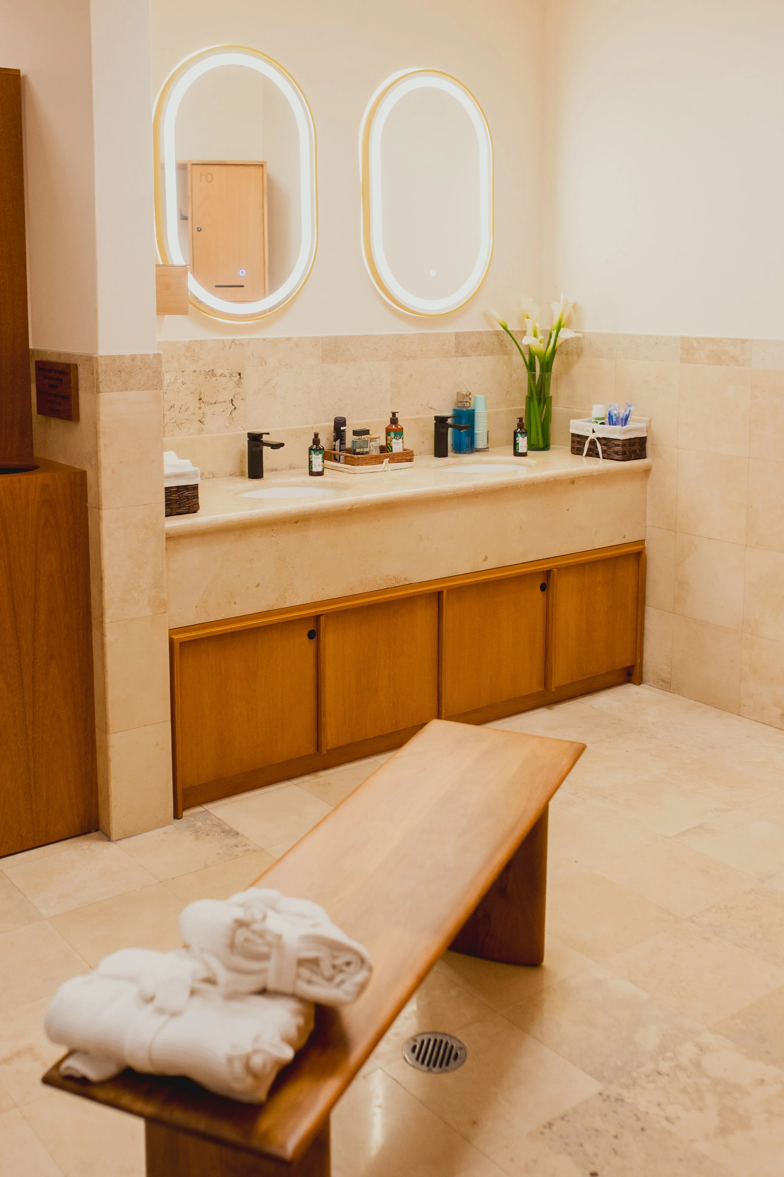 A bathroom sink area with two oval mirrors, a beige marble countertop, a wooden cabinet below, and a bench with folded towels. There are soap dispensers, a small tray of toiletries, a tall vase with white flowers, and a basket with toothbrushes on th