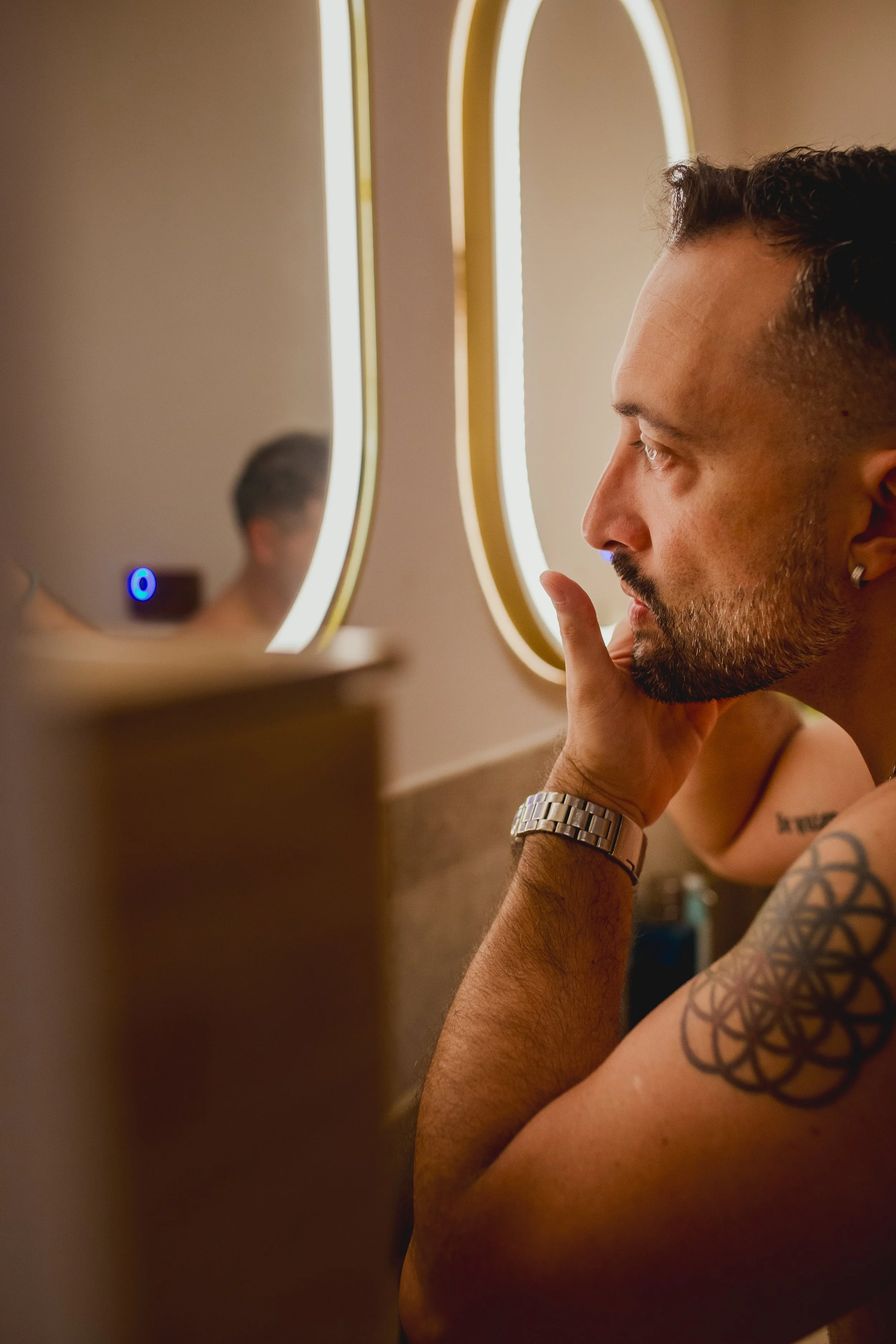 A man with tattoos and a wristwatch looks into a mirror while touching his chin, in a bathroom with circular backlit mirrors.
