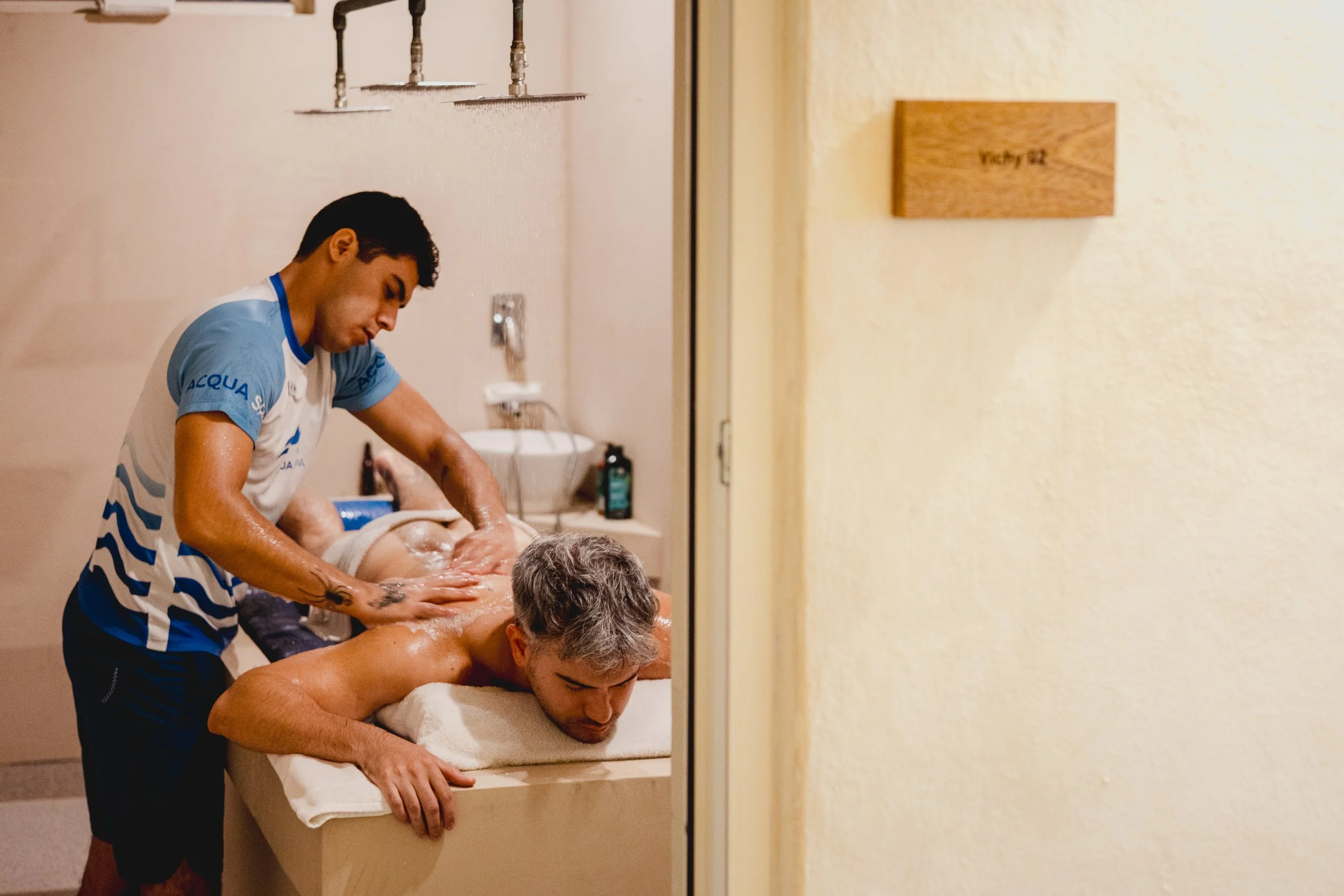 A man giving a massage to another man who is lying face down on a massage table. The person receiving the massage is covered with a towel and is outdoors or in a room with a visible door or partition.