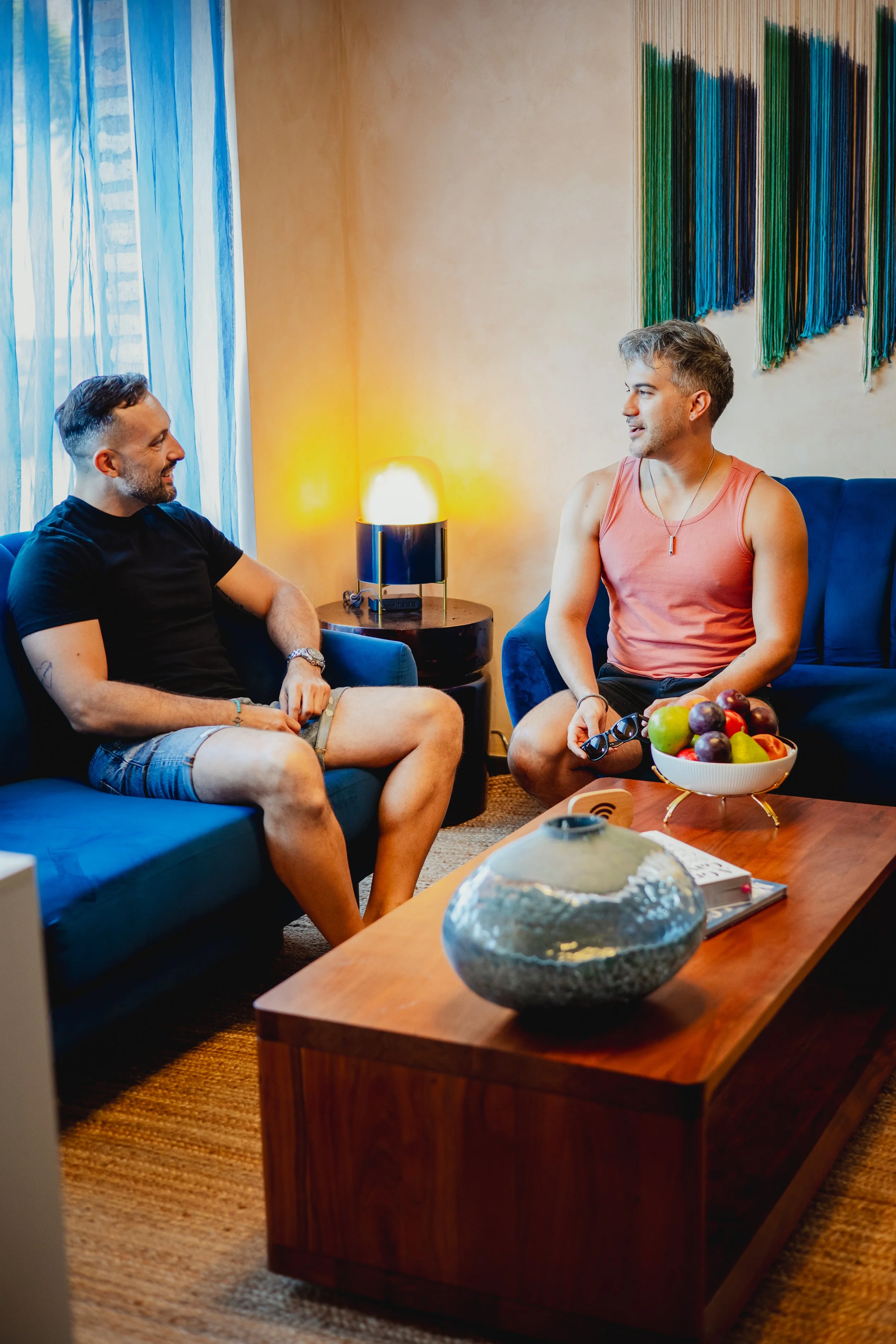 Two men are sitting on blue couches in a living room, engaged in conversation. One man is wearing a black t-shirt and shorts, the other is in a sleeveless orange top and shorts. There is a wooden coffee table with a bowl of fruit and a decorative vas