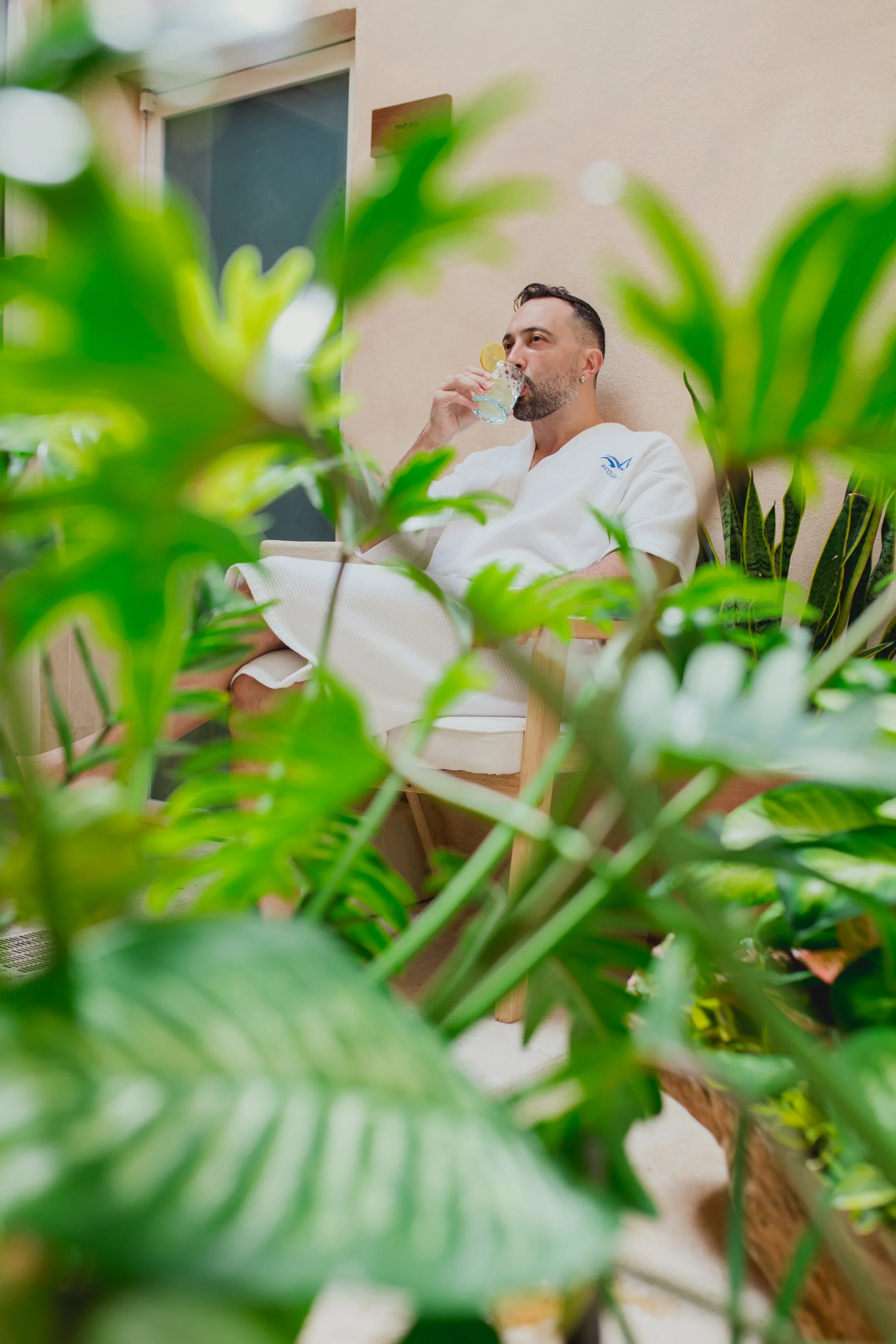 Man in a white bathrobe sitting on a chair, drinking water with lemon slices, surrounded by lush green plants indoors.