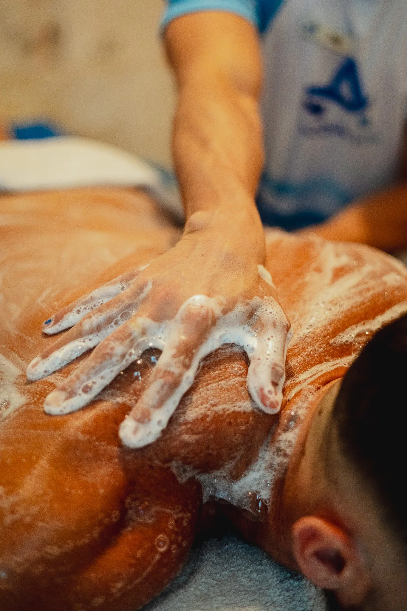 Person receiving a massage, lying face down, with soap suds on back, while massage therapist applies pressure with hand.