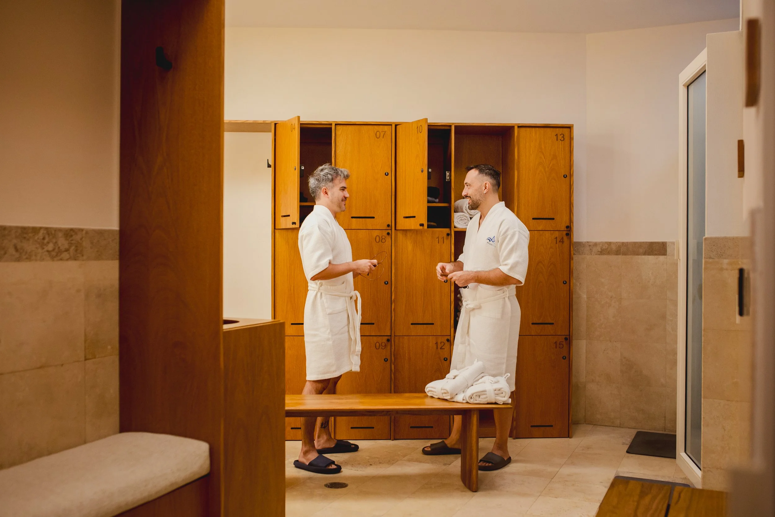 Two men in white bathrobes are talking in a locker room with wooden lockers and a wooden bench, wearing slippers and holding towels.