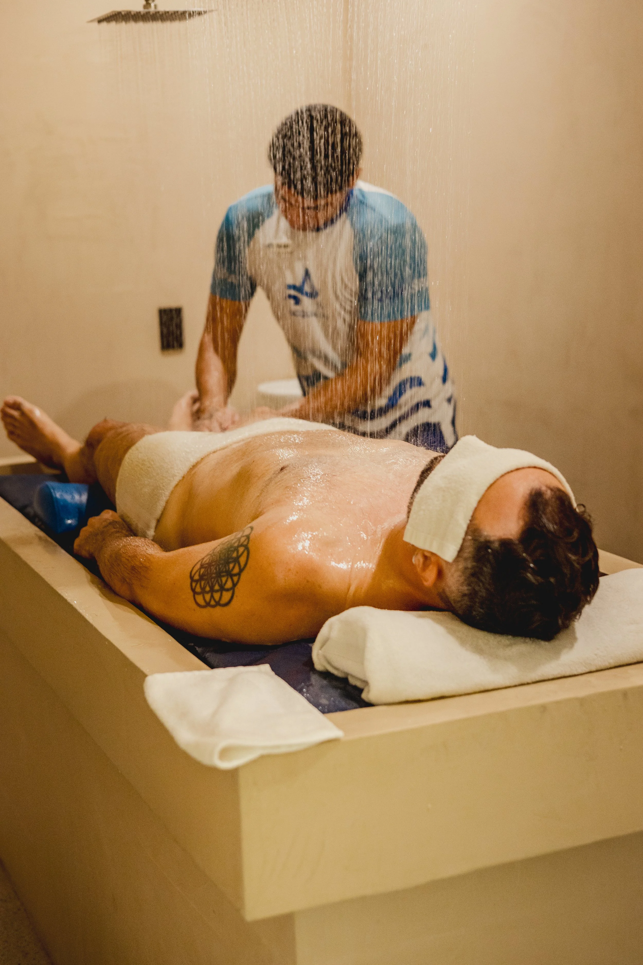 A man receiving a massage in a spa or massage room, lying face down on a massage table, covered with a towel, while a therapist washes him under a rainfall shower head.