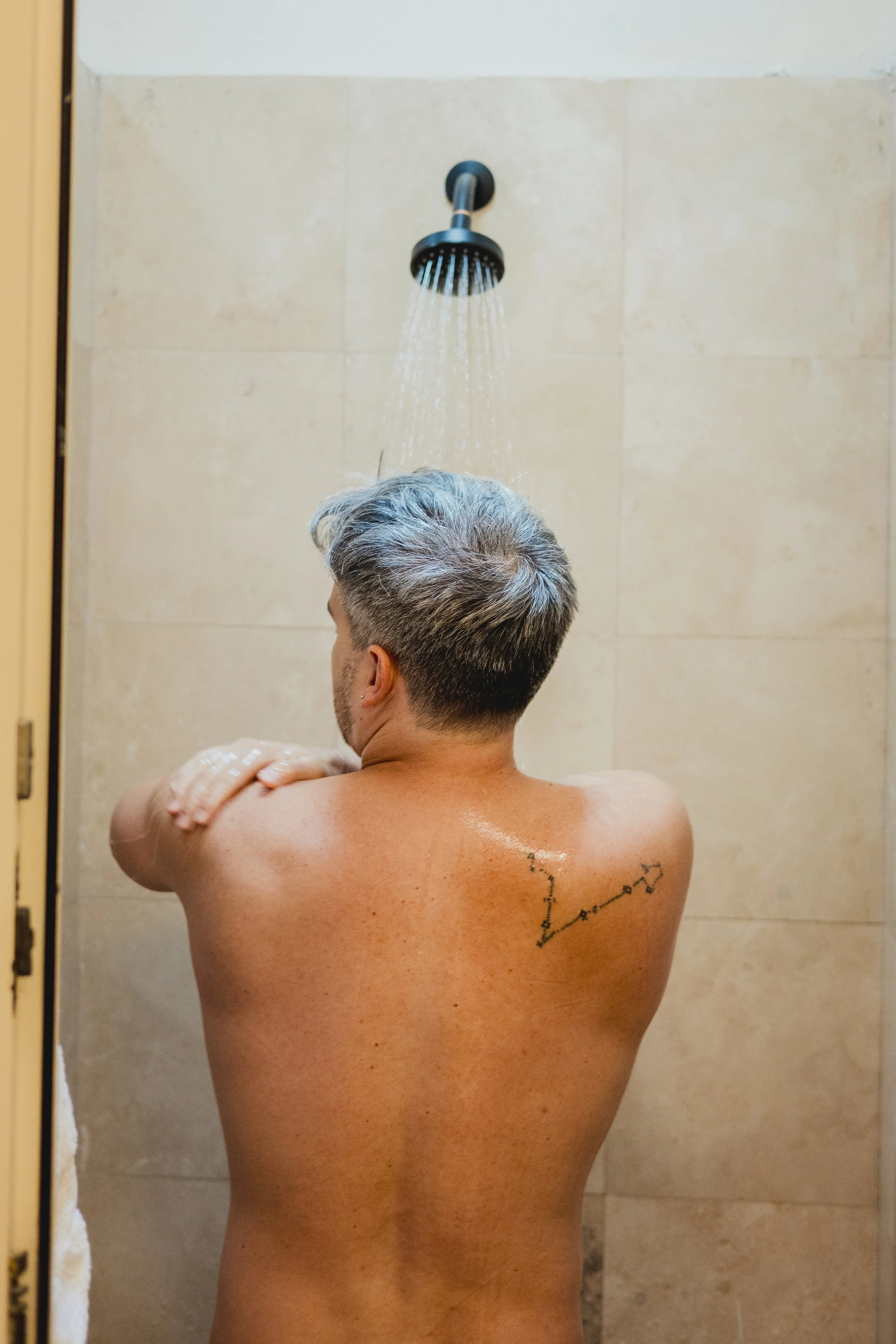 A man with gray hair taking a shower in a bathroom with beige tiled walls, wearing no clothes, with a tattoo on his upper back.