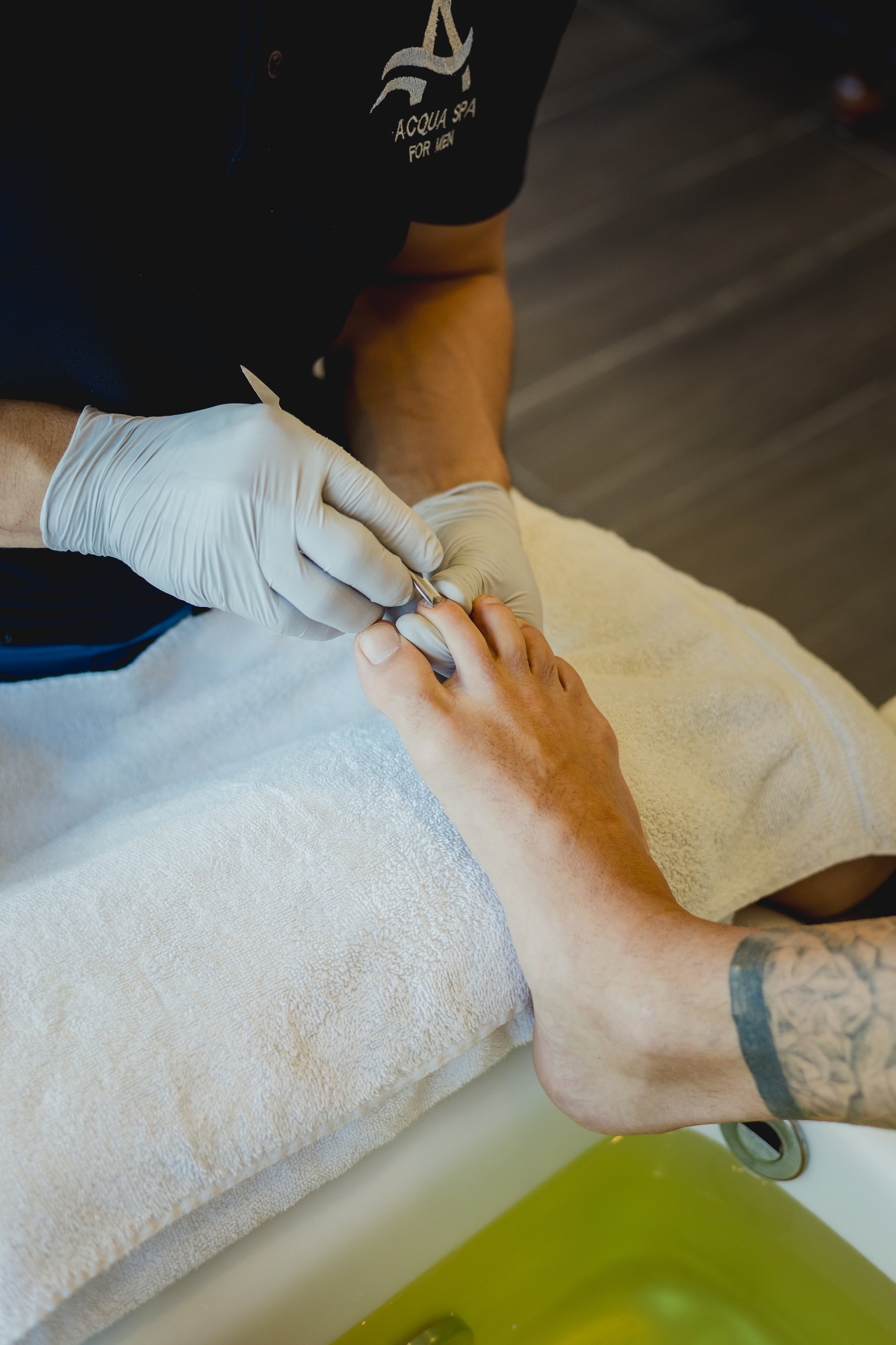 A person receiving a professional pedicure at a spa, with a nail technician filling a client's toenail with a manicure tool, while the client's foot rests over a basin of water.