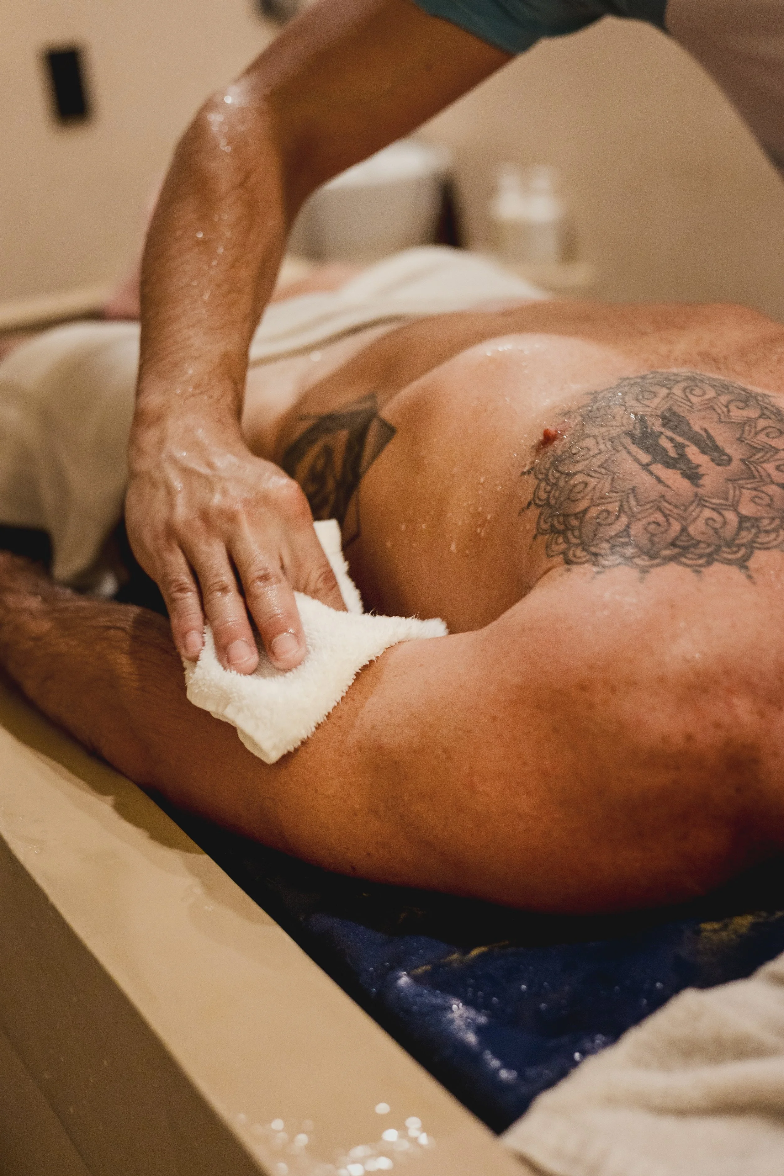 A person receiving a massage, lying on a massage table with a towel and some water droplets on their back, and a tattoo on their shoulder blade.