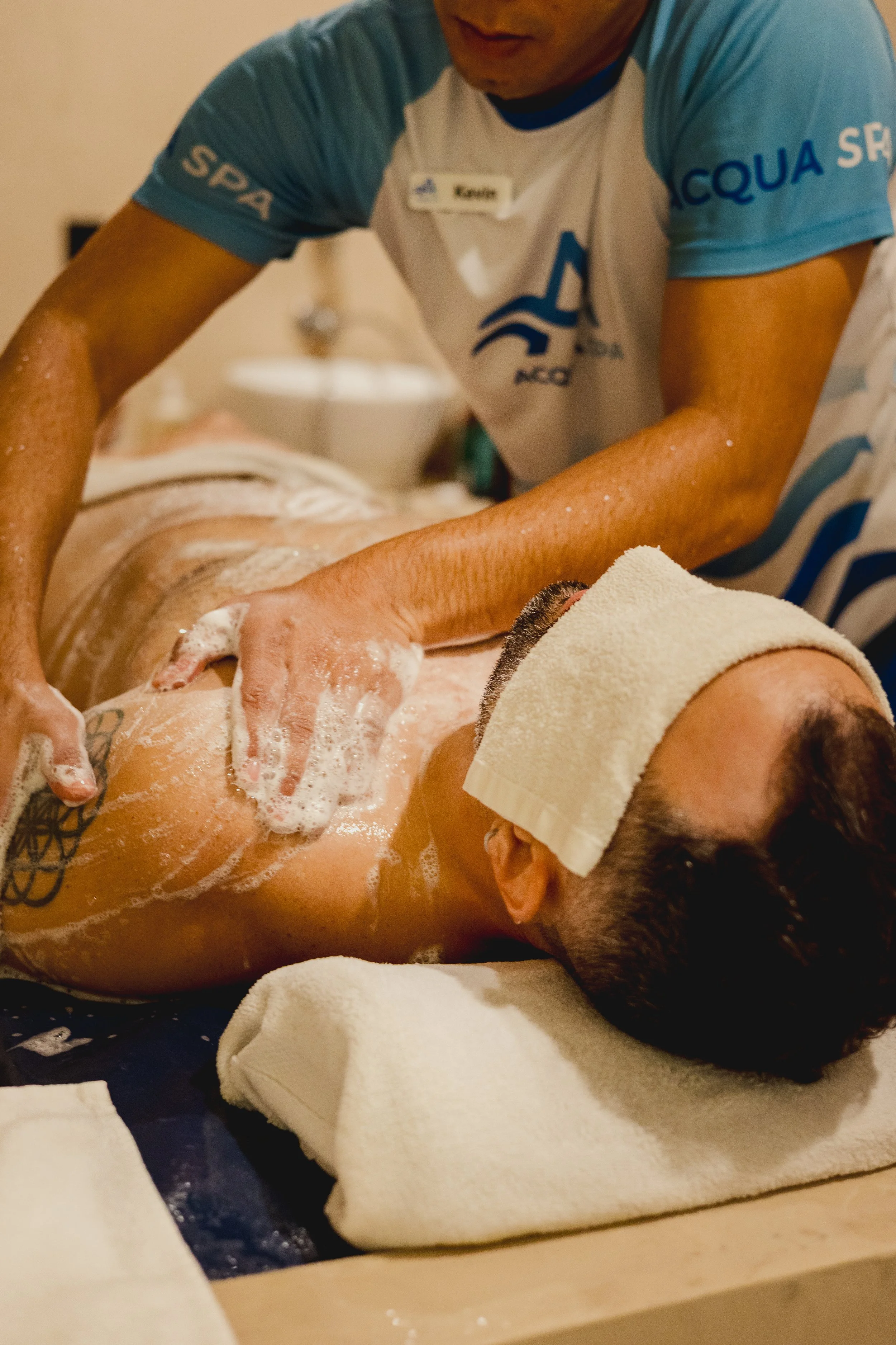 A person is receiving a massage therapy with soap and foam on their back, lying face down on a massage bed. The therapist is wearing a name tag and a blue and white shirt.