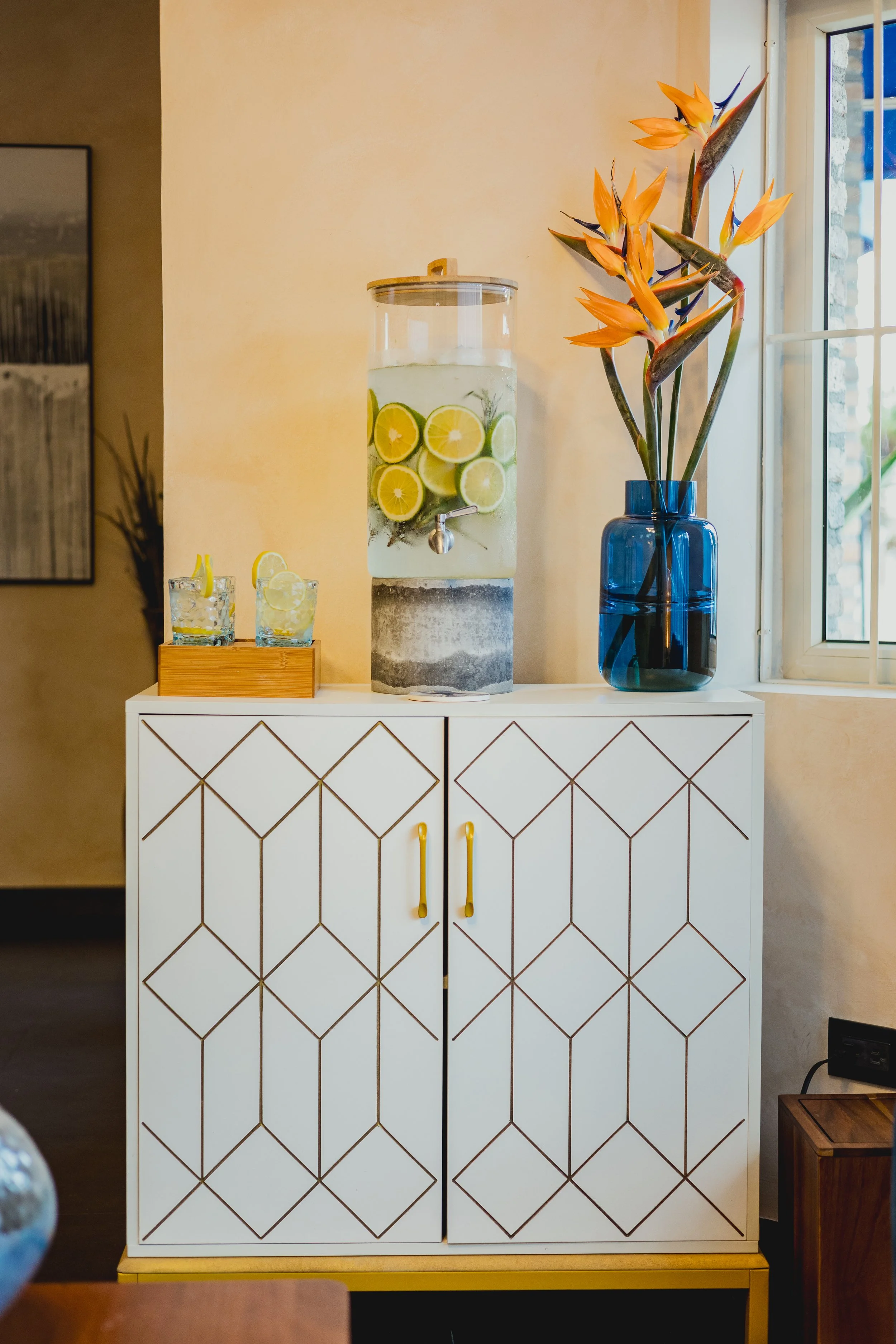 A white cabinet with geometric patterns, decorated with a large glass water dispenser filled with lemon and lime slices, a blue vase with orange flowers, and two glasses of lemon water on a tray, near a window in a cozy room.
