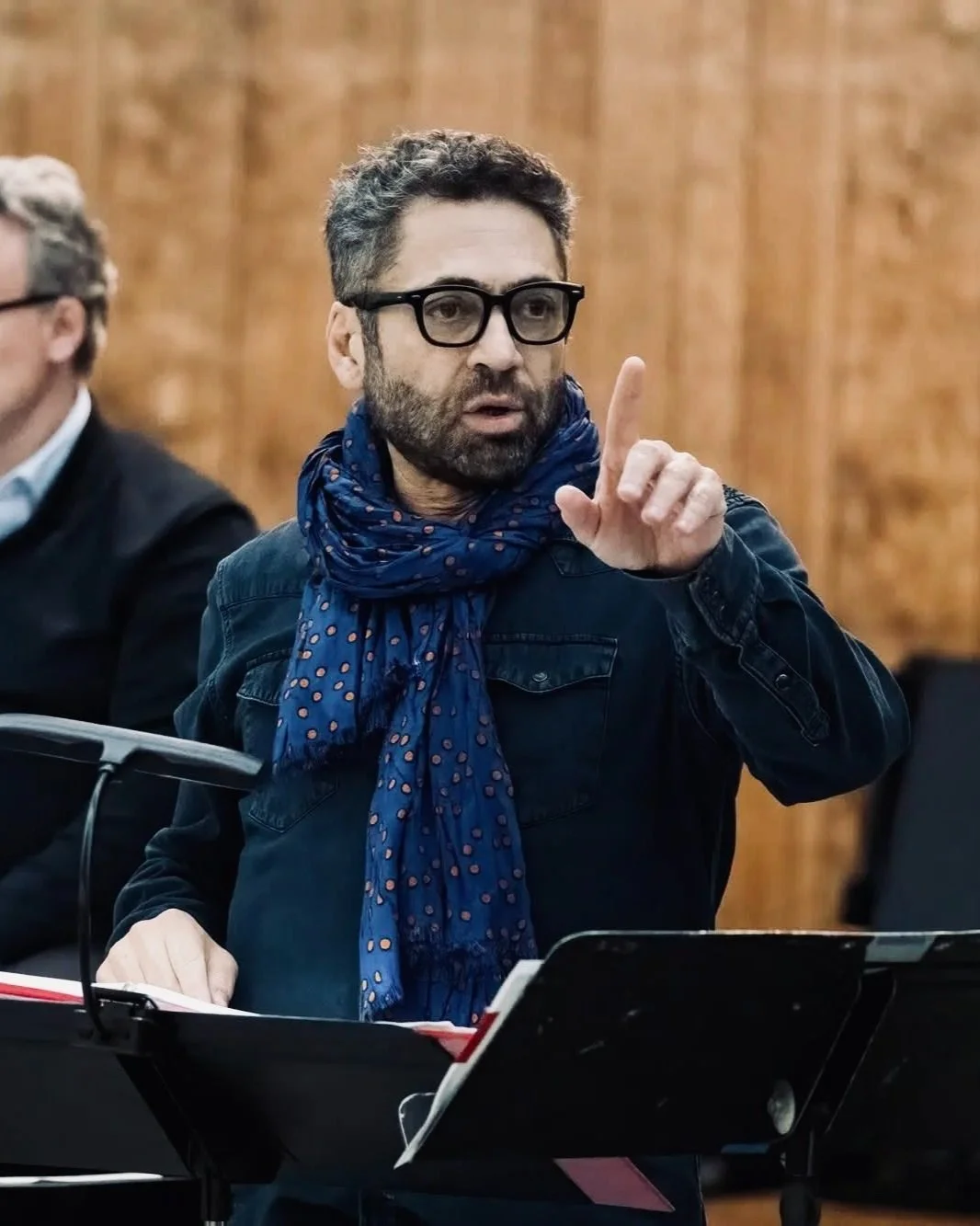Man wearing glasses and a blue scarf, speaking with an expressive gesture, standing in front of music stands indoors.