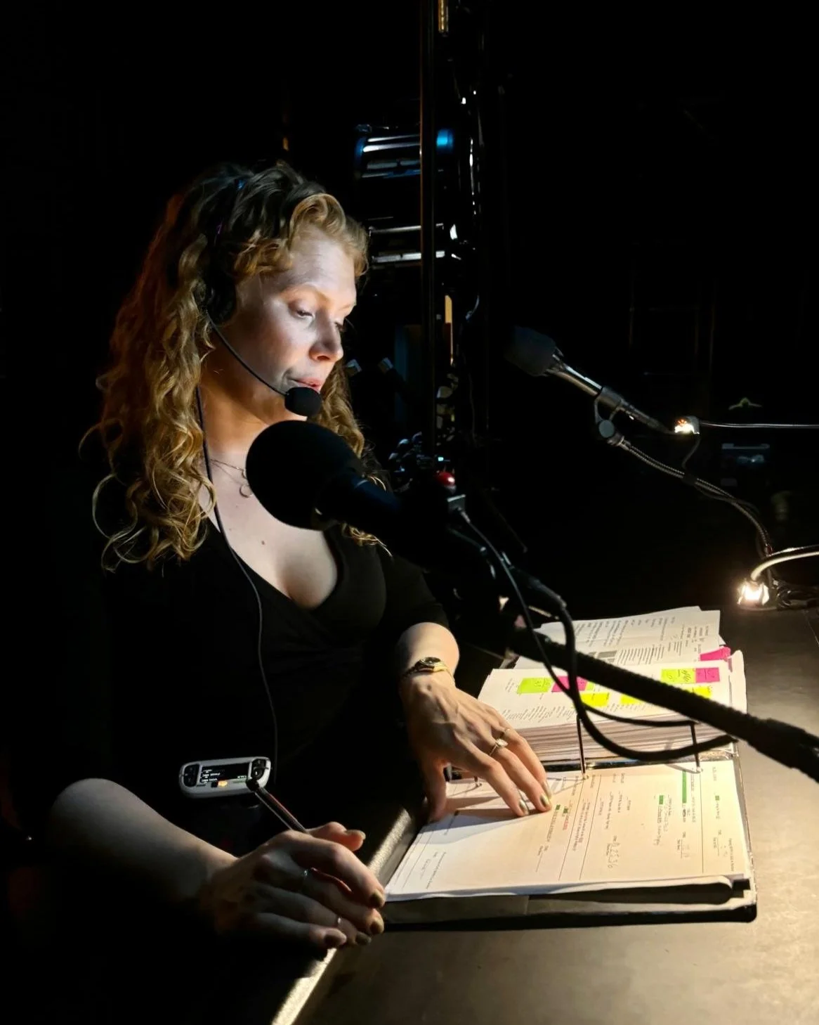 A woman wearing a headset, sitting at a desk with a microphone, reading from a script with highlighted notes.