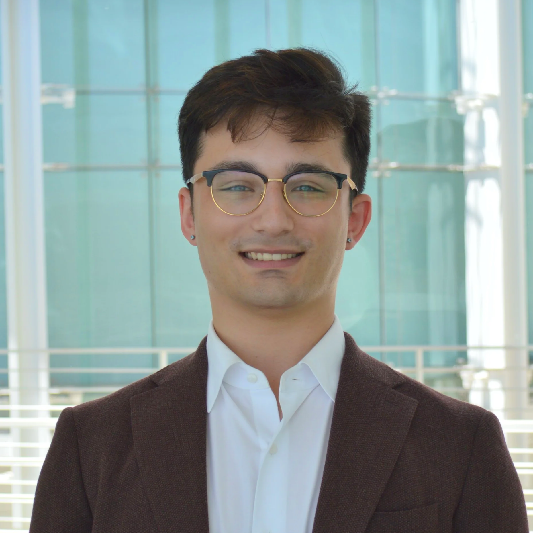 A young man with dark hair, wearing glasses, a white shirt, and a brown blazer, smiling in front of the city hall rotunda..