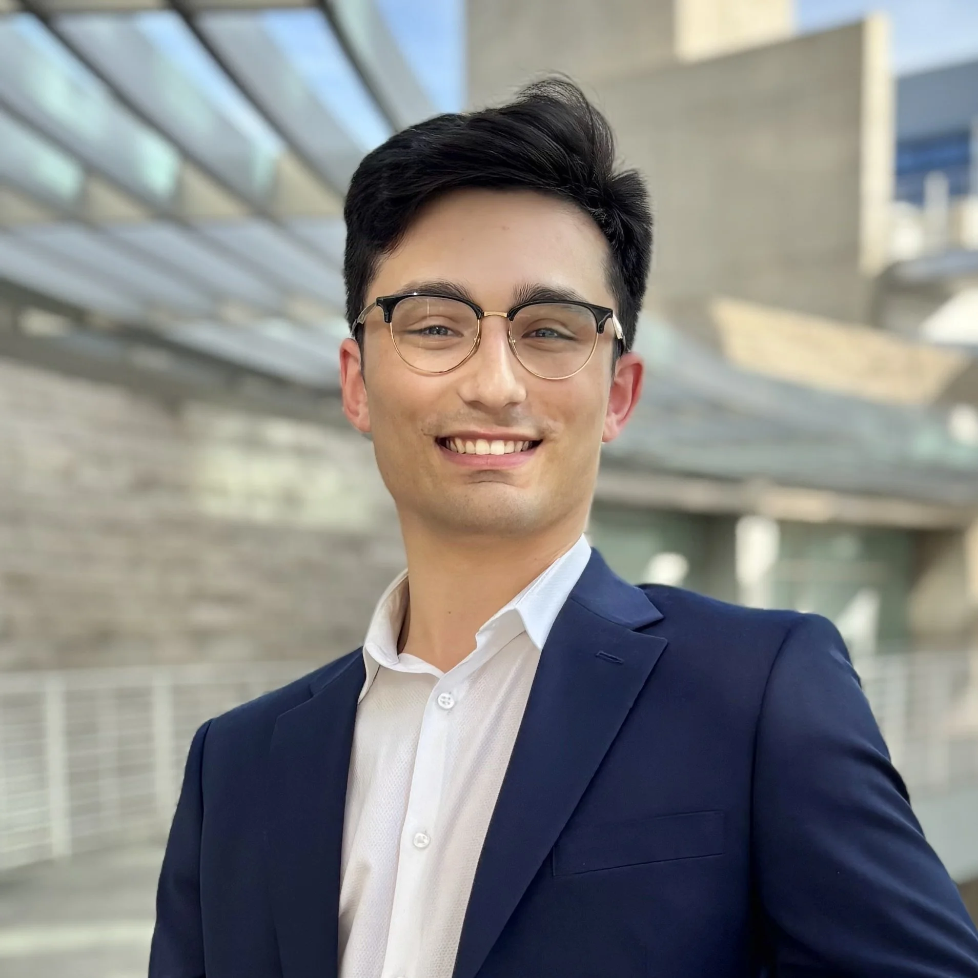 A young man with dark hair, wearing glasses, a white shirt, and a navy blazer, smiling against a modern office background.