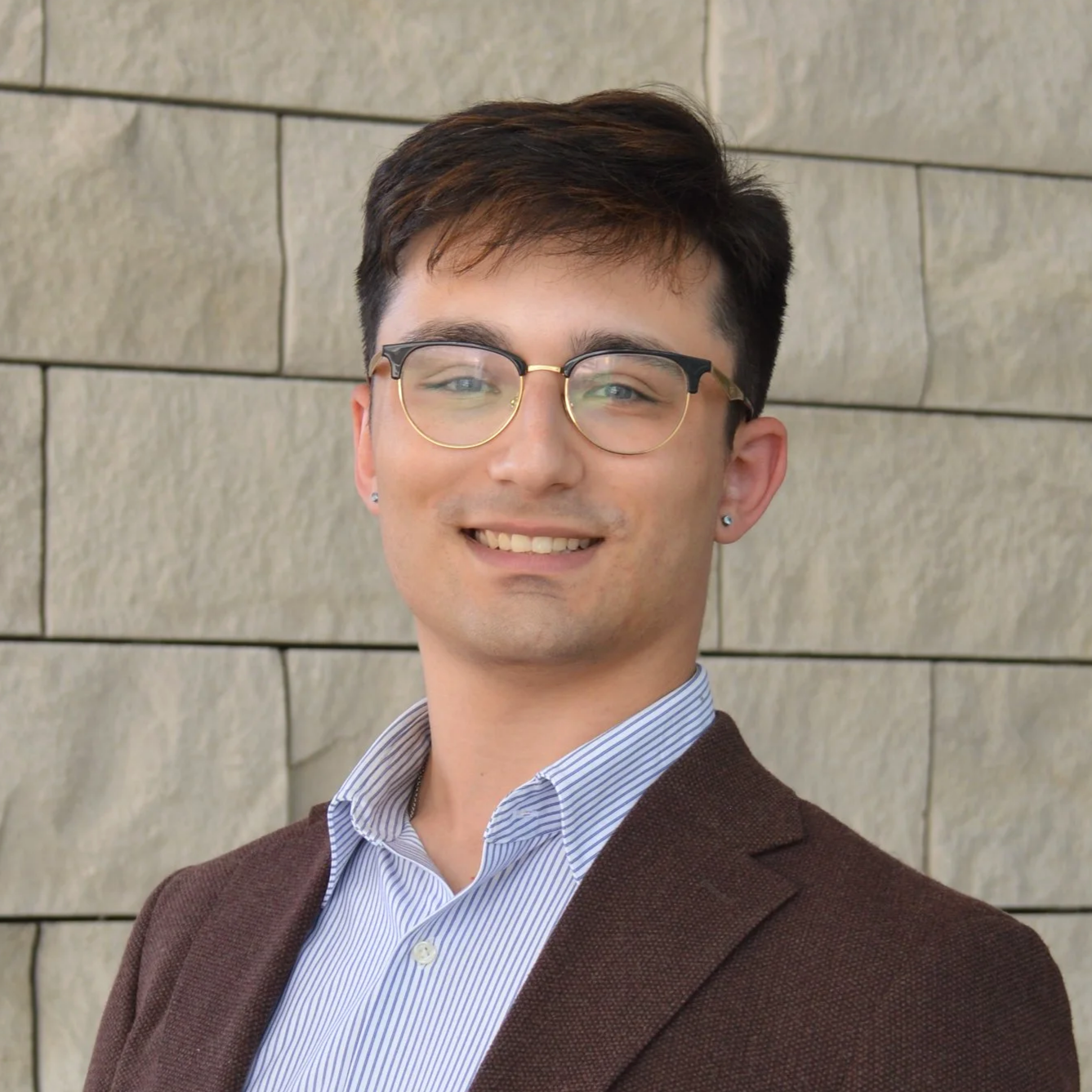 A young man with dark hair, wearing glasses, a white shirt, and a brown blazer, smiling in front of the city hall rotunda..
