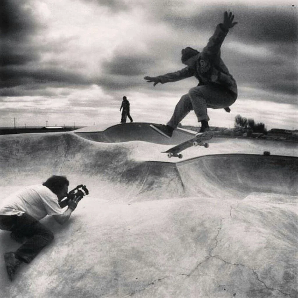 John Reeves aka JTMR does a 1-foot Ollie over the hip of Browning Skatepark, on the Blackfoot Reservation in Montana. While Oscar Wagenbulcher films on a video camera  and another person stands in the background under cloudy skies.