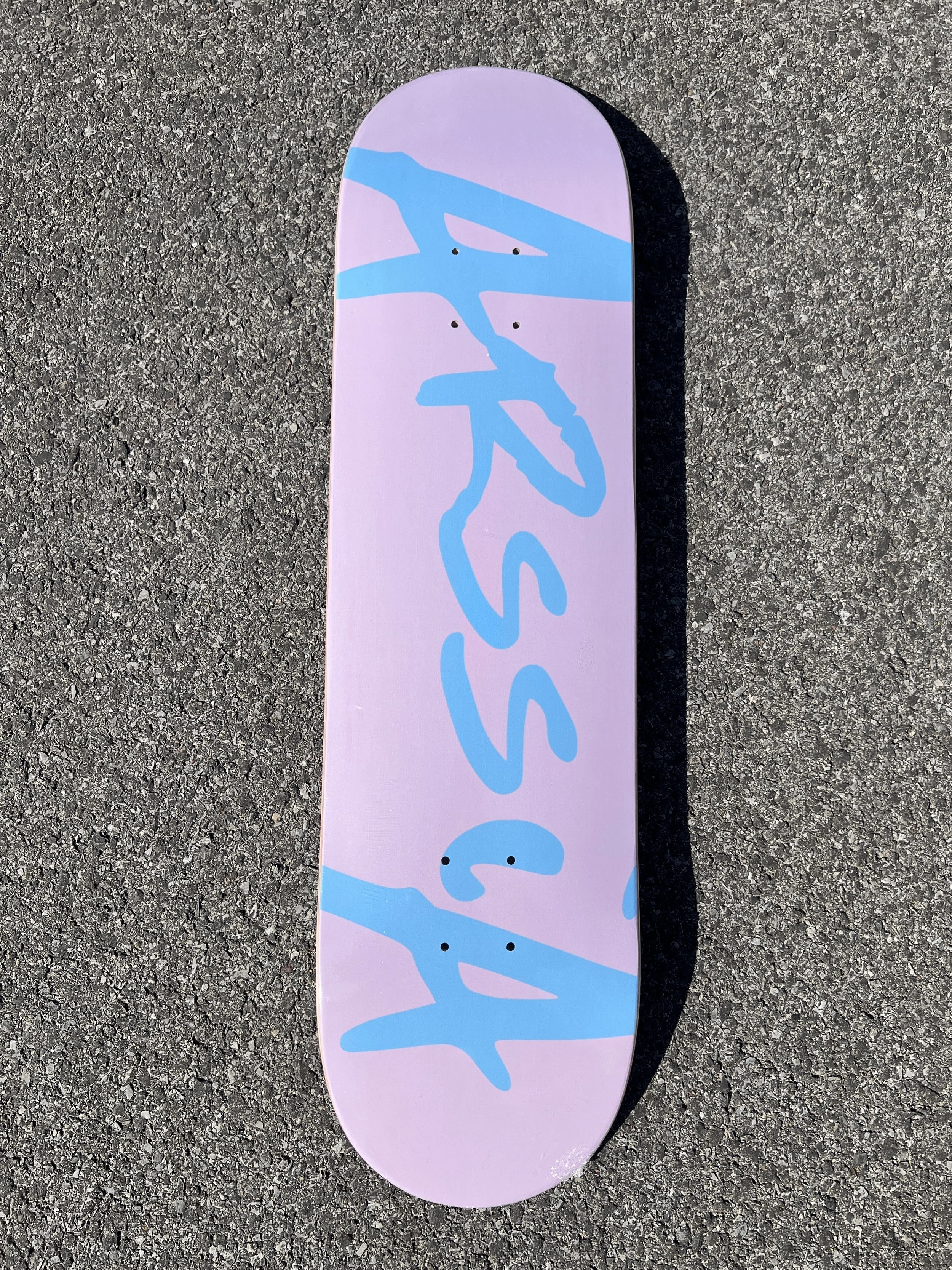 ArssiA- “O.G. Logo” skateboard with a pink deck and blue graffiti-style text and design, lying on asphalt.