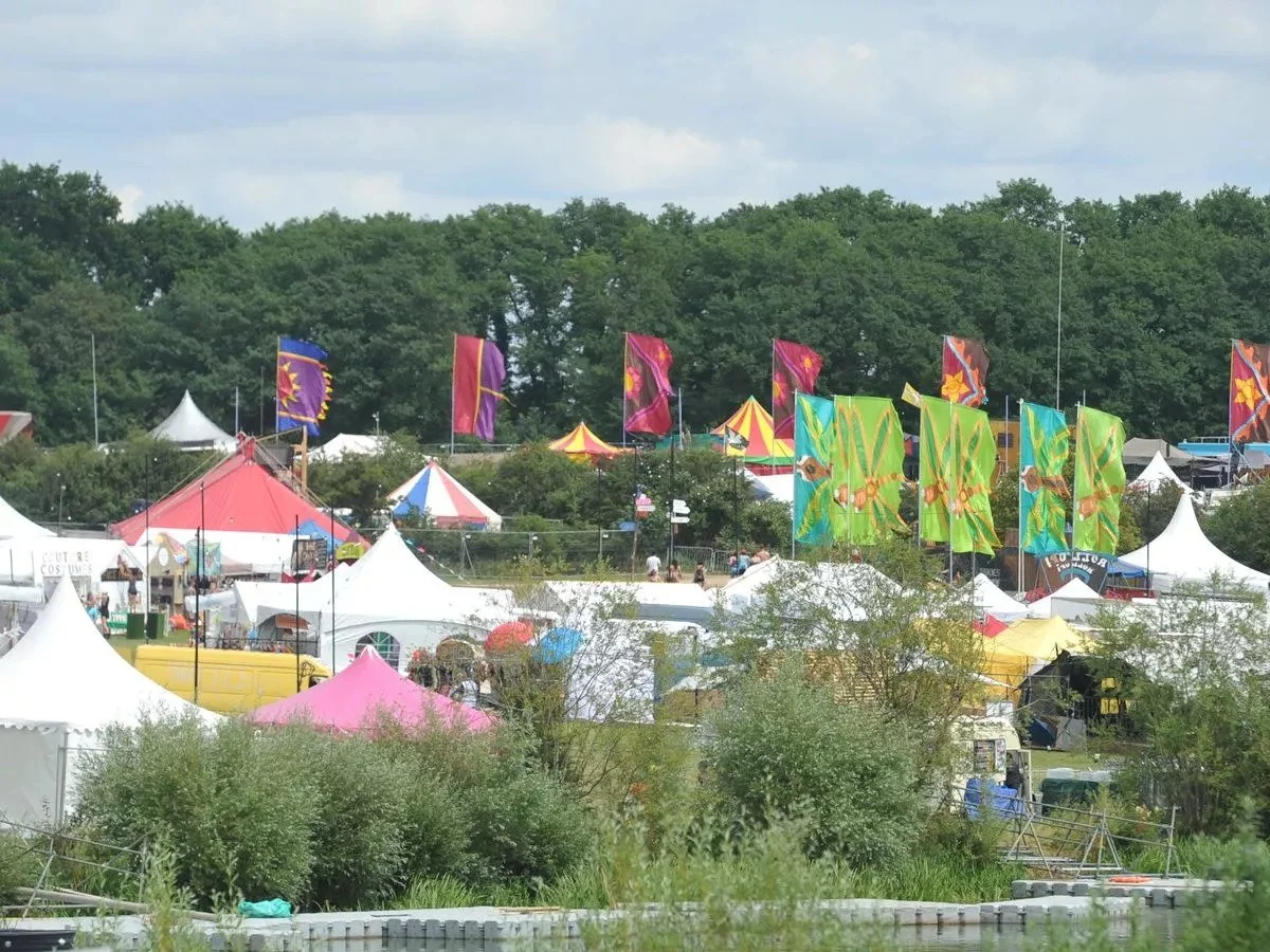 A colorful fair with various tents, flags, and banners amidst trees during the day.