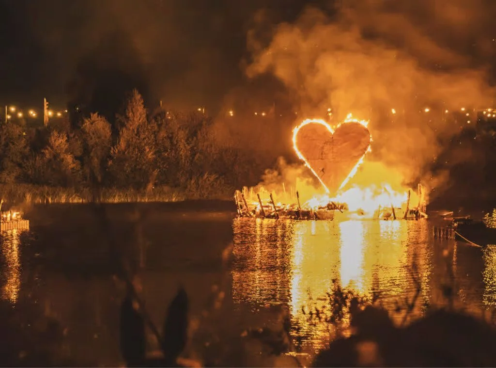 A large fire on a body of water, with a heart shape outlined in flames above the burning wreckage.