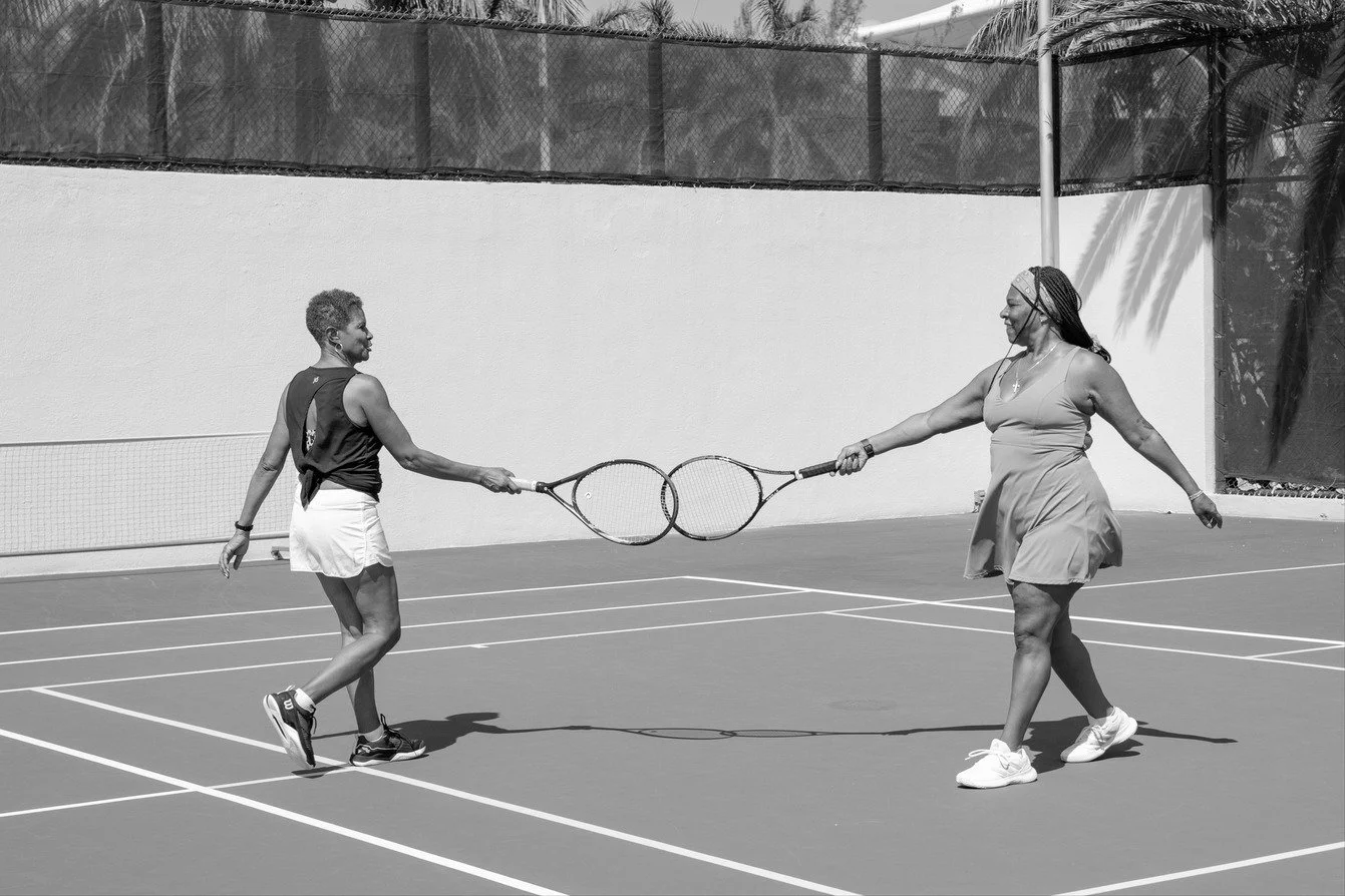 Two women playing tennis on an outdoor court with tennis rackets, sitting and smiling at each other.