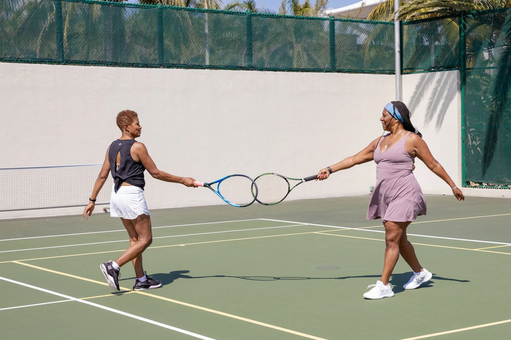 Two women playing tennis and engaging in a playful sword fight with tennis rackets on an outdoor tennis court.
