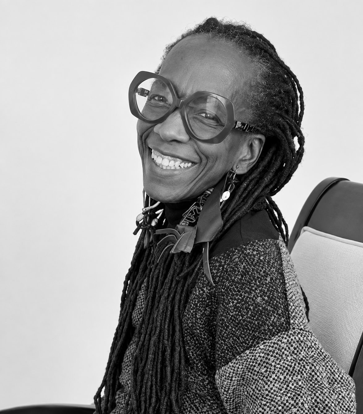 A woman with long hair in dreadlocks wearing large glasses, earrings, and a patterned jacket, smiling while sitting on a chair.