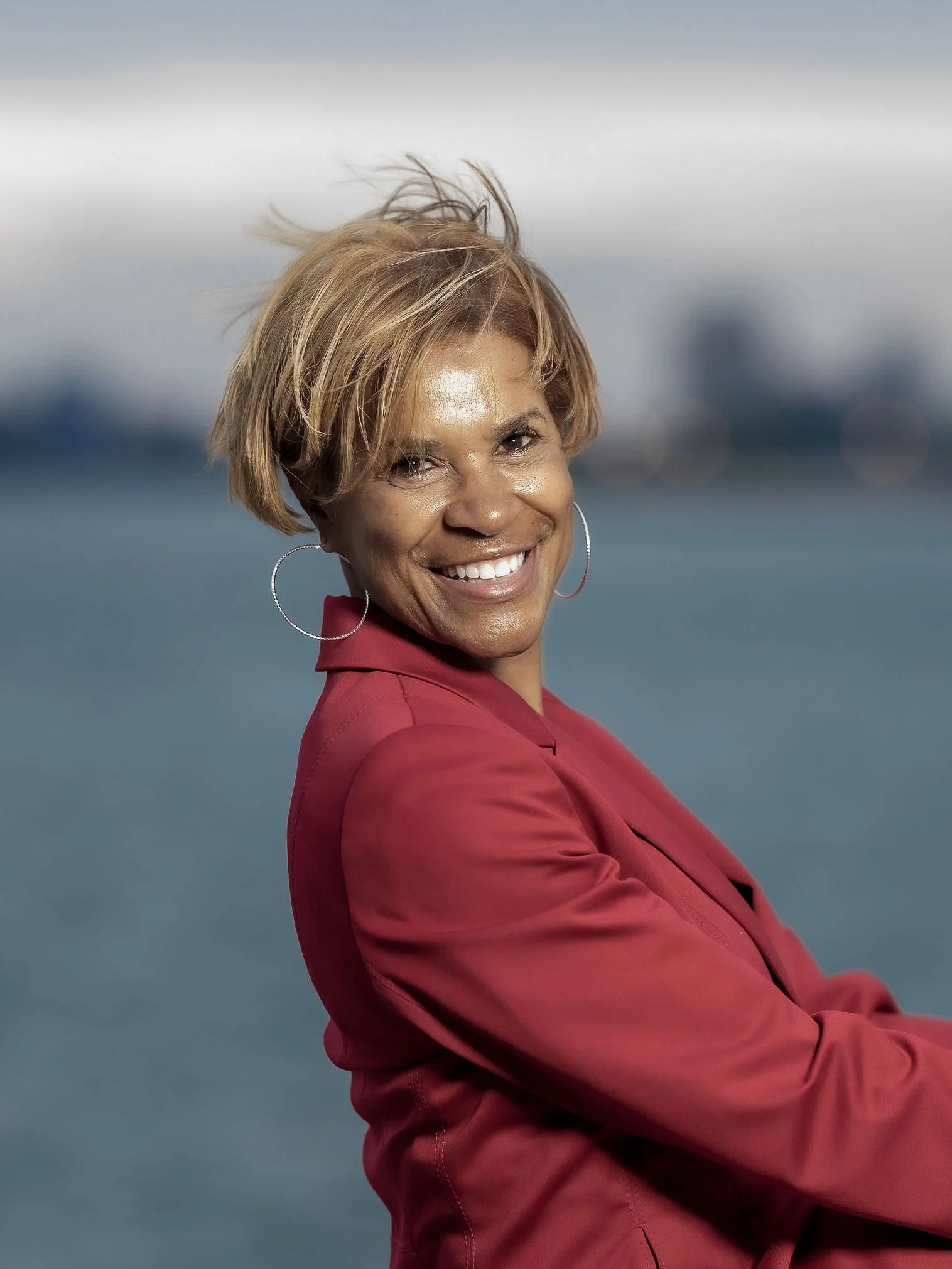 A smiling woman with short blond hair, wearing hoop earrings and a red blazer, standing outdoors with a blurred cityscape and sky in the background.