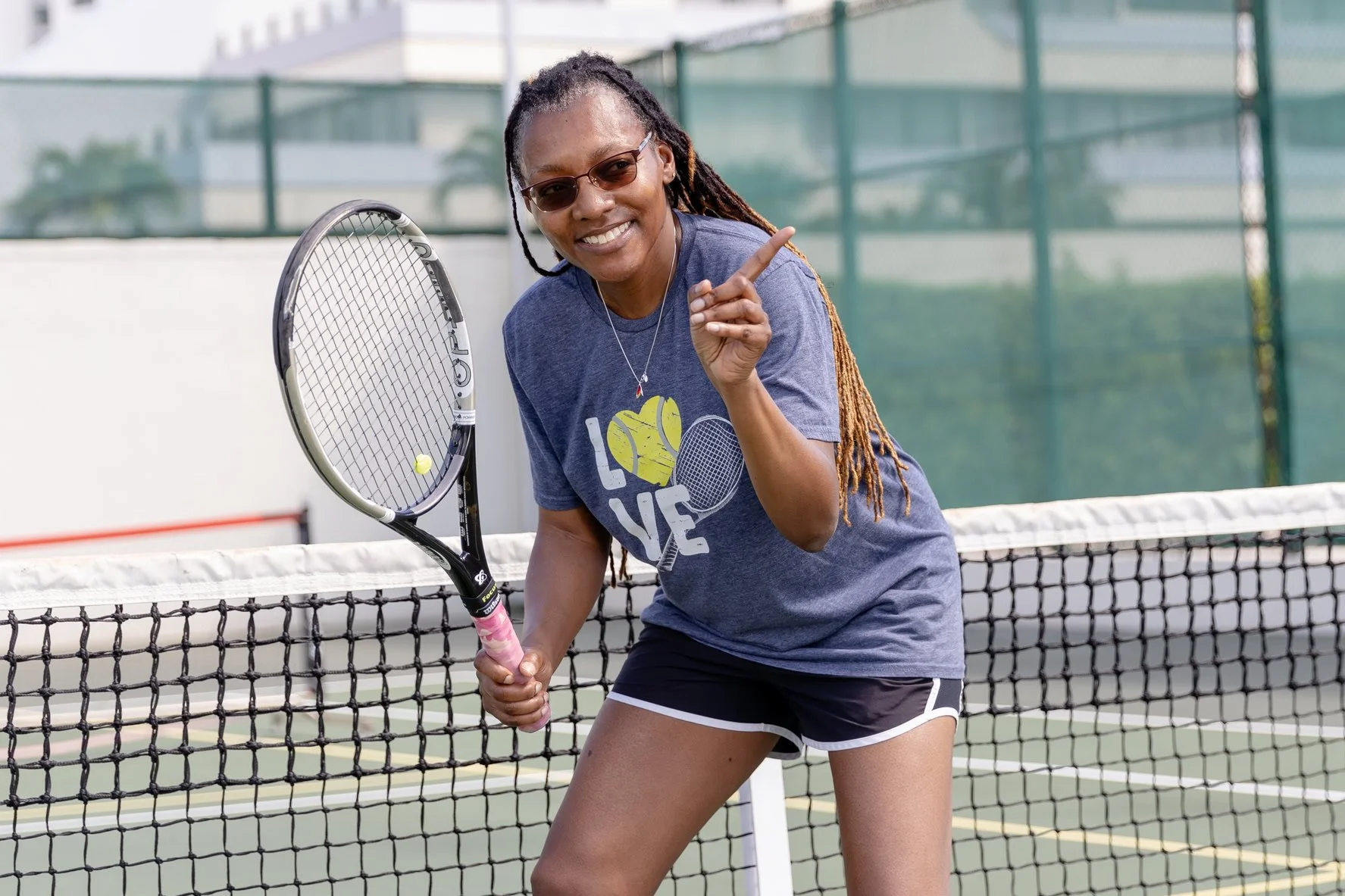A woman with long dreadlocks and sunglasses on a tennis court, smiling and pointing with her index finger, holding a tennis racket, with a tennis ball nearby.