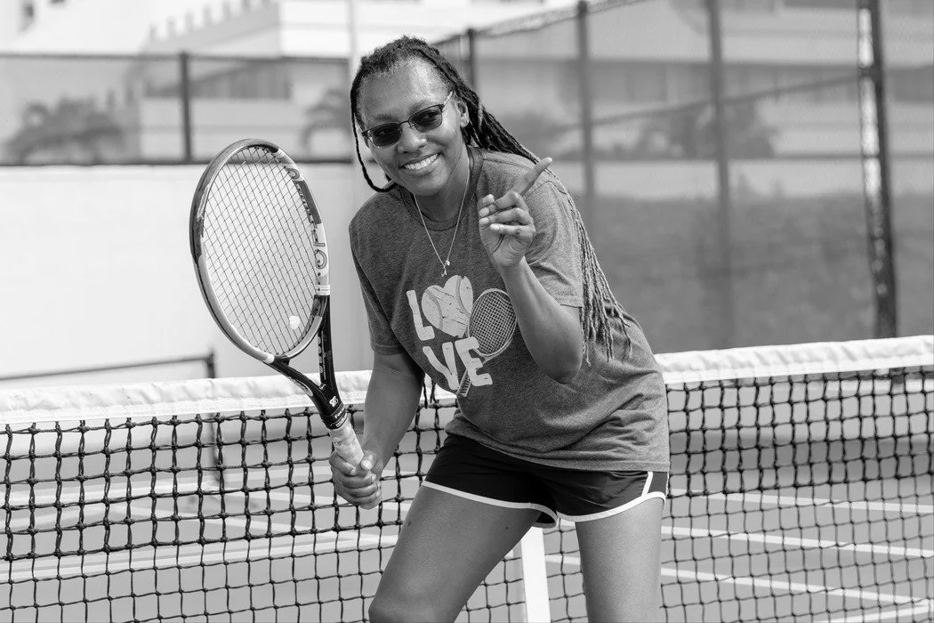 A woman with dreadlocks wearing sunglasses, a T-shirt and shorts, holding a tennis racket on a tennis court with a net, smiling and pointing with her finger.