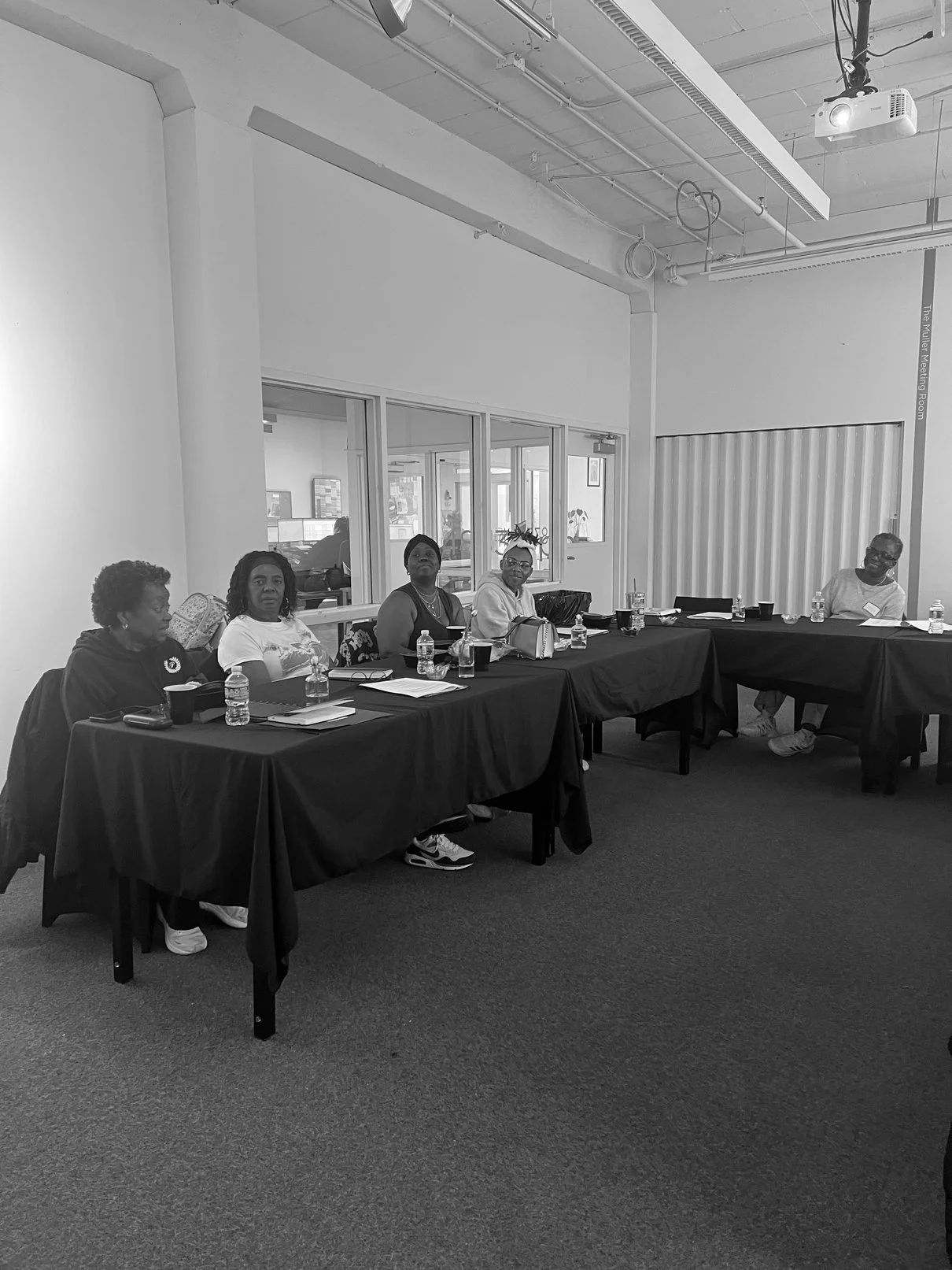 A group of five women sitting at a long black cloth-covered table, with water bottles, notebooks, and cups in front of them, in a room with a high ceiling and a projector mounted above.
