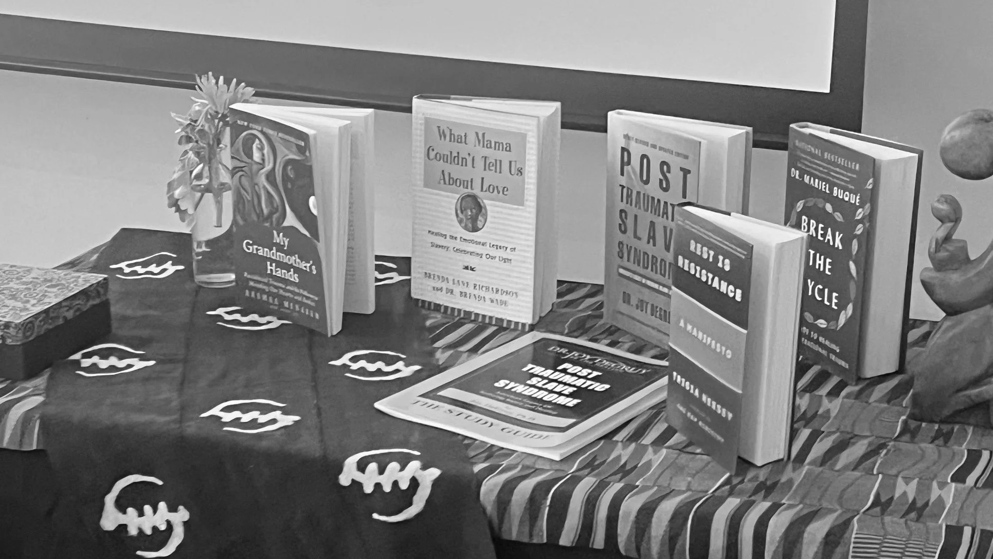 Display of several books related to trauma, love, and mental health on a table with a Halloween-themed tablecloth, alongside a vase with flowers and flyers about post-traumatic slave syndrome.