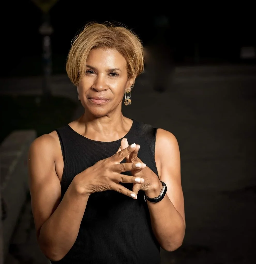 A middle-aged woman with short, blonde hair standing outdoors at night, wearing a black sleeveless top, with her hands clasped together, looking directly at the camera.