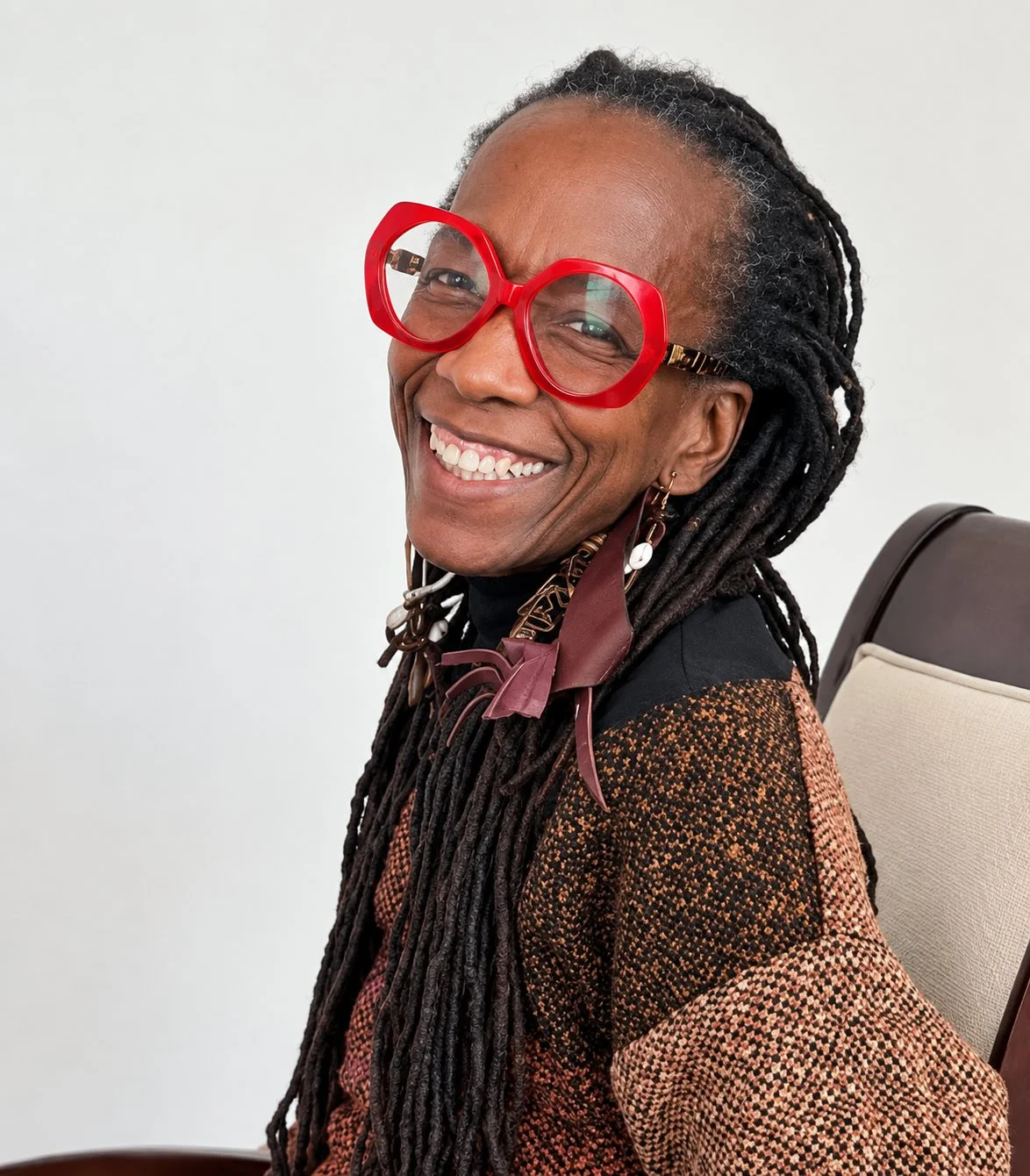 A smiling woman with glasses, dreadlocks, and earrings seated on a beige chair against a plain white background.