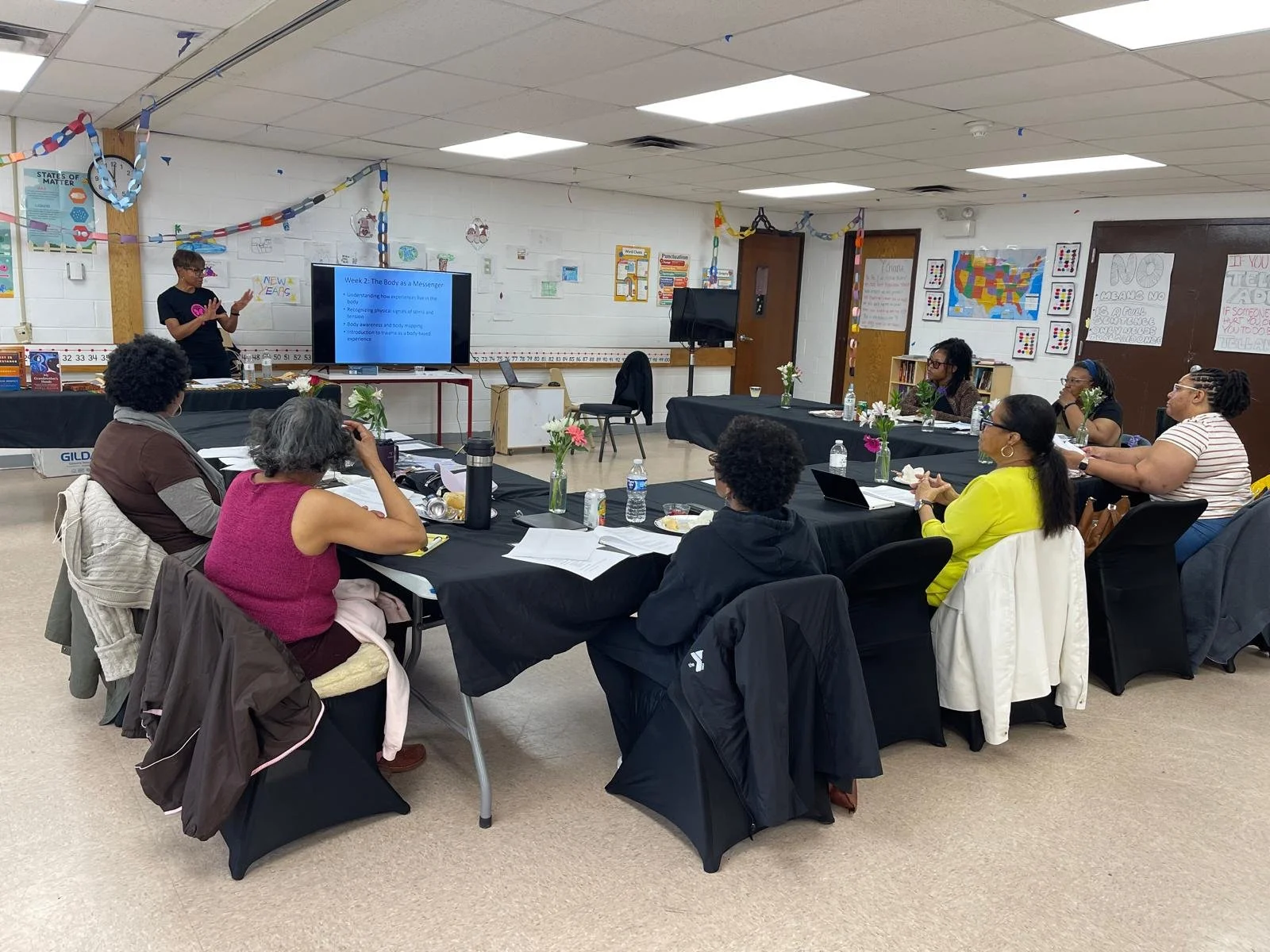 A classroom or workshop setting with a group of women sitting around tables listening to a woman giving a presentation. There is a large screen displaying presentation slides, with cups, water bottles, and documents on the tables. The background features educational posters and decorations, with some colorful chains hanging from the ceiling.