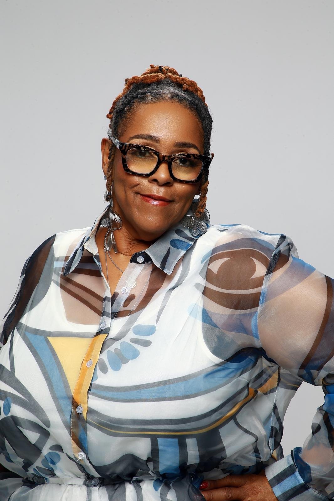 A woman with glasses and earrings, wearing a colorful patterned blouse, smiling and posing with her hand on her hip against a plain light background.