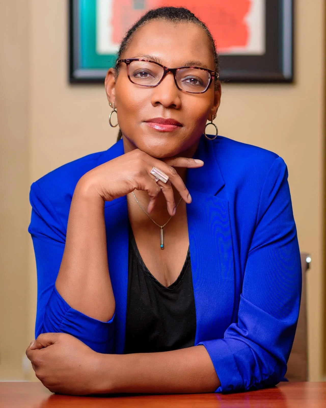 A woman wearing glasses, a blue blazer, and jewelry, sitting at a desk with her chin resting on her hand, in front of a blurred background with a framed picture.