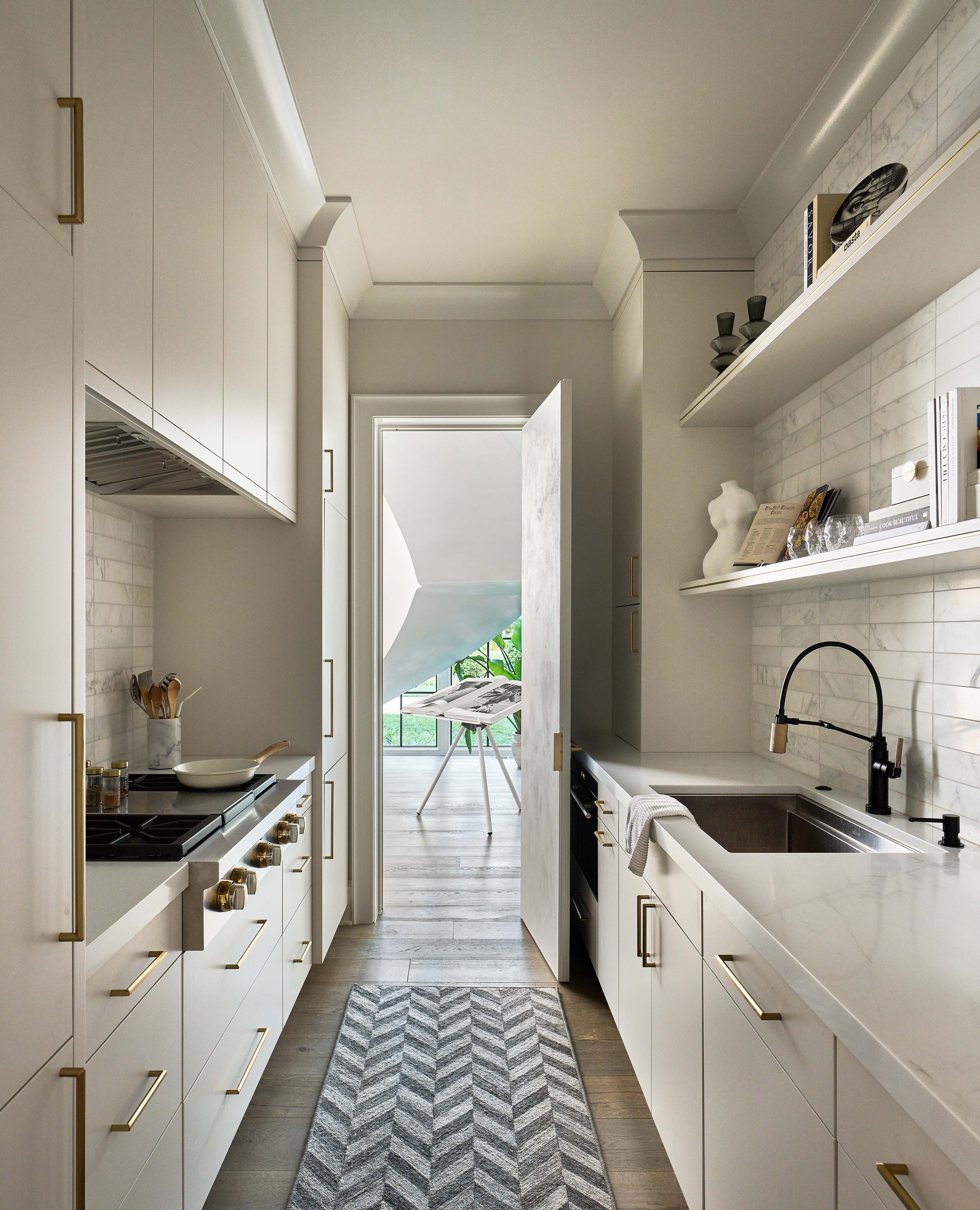 Modern kitchen with white cabinets, gold handles, black faucet, chevron rug, and open door leading to an outdoor seating area.